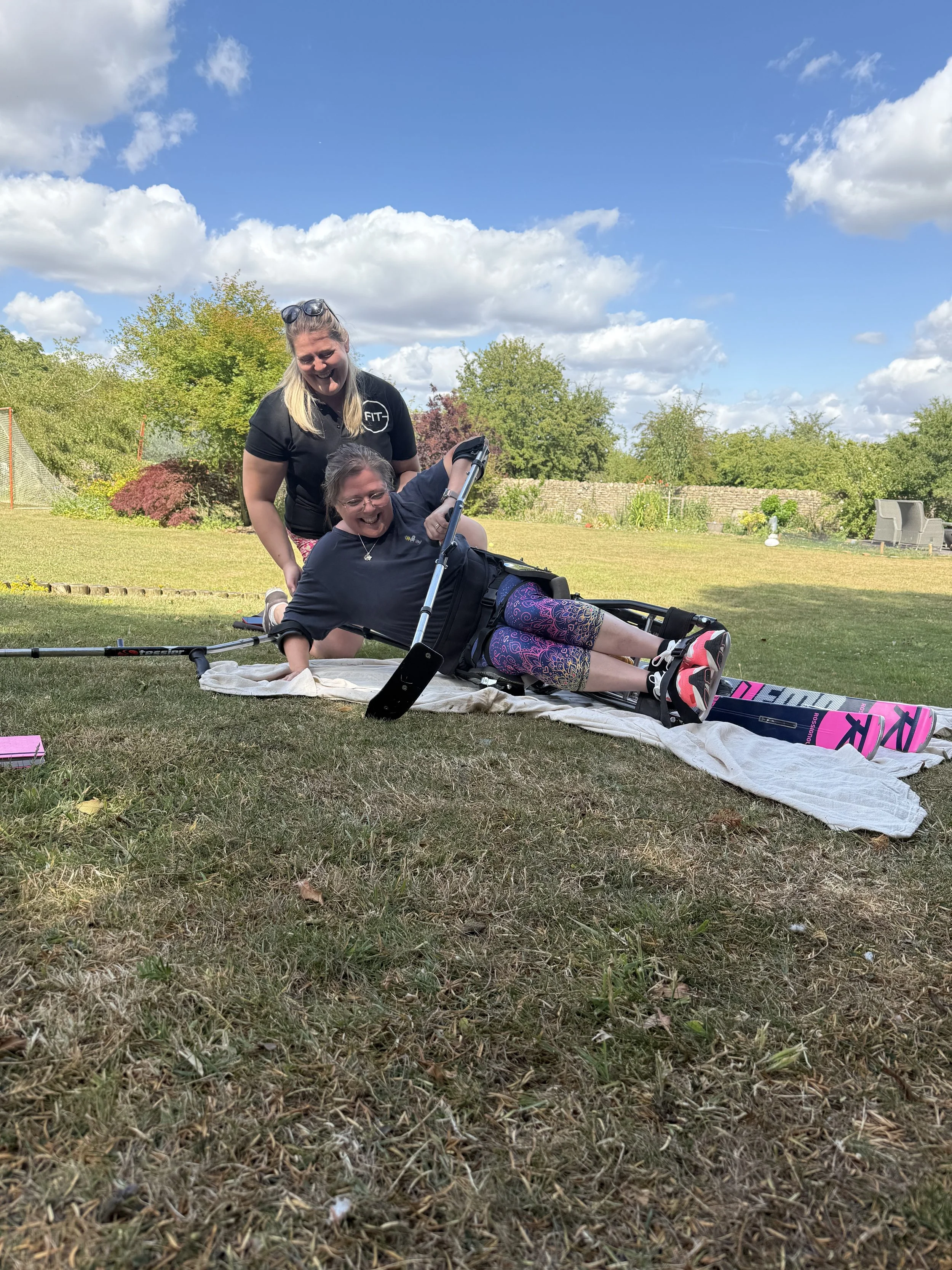 Two women outdoors on a grassy lawn, one assisting the other who is lying on the ground beside a wheelchair, with a bright blue sky and trees in the background.