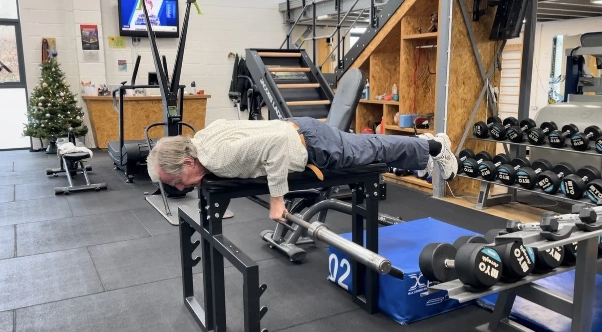 A man performs a seal row exercise while lying on a padded bench and legs extended behind him in a gym, surrounded by weights and exercise equipment.