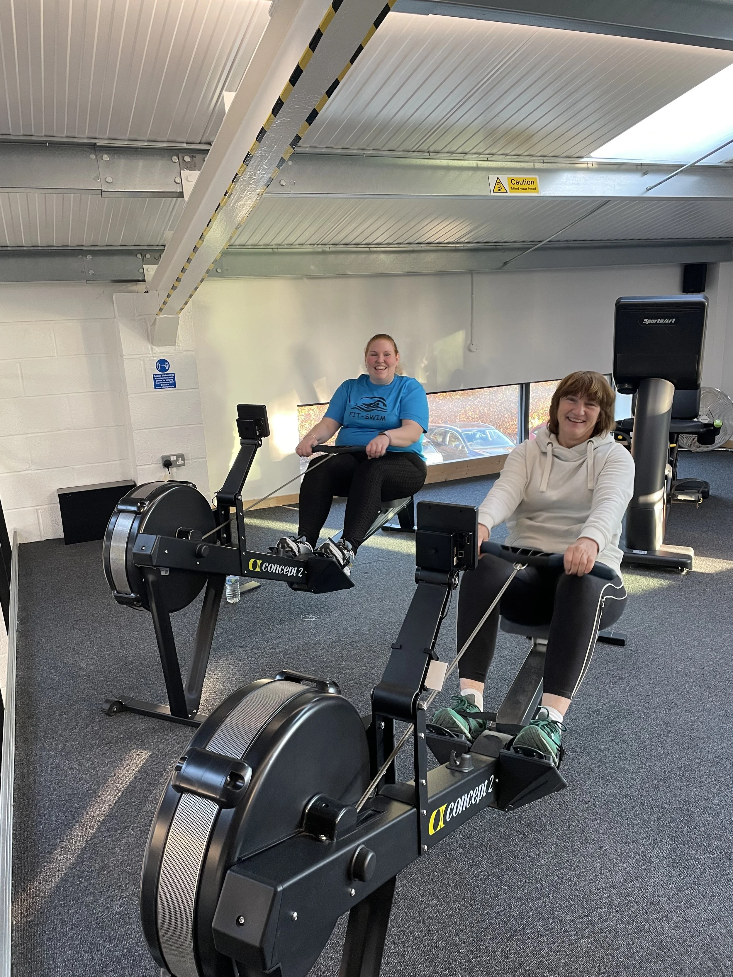 Two women are exercising on rowing machines inside a gym. They are smiling and appear to be enjoying their workout. The gym has a gray carpeted floor, a white brick wall, and large windows that let in natural light. There is a water bottle on the floor near one of the machines.