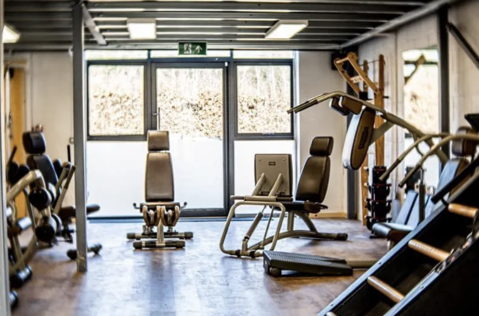 Empty gym room with various workout equipment including exercise chairs and weight machines, large windows, and an exit sign above the door.