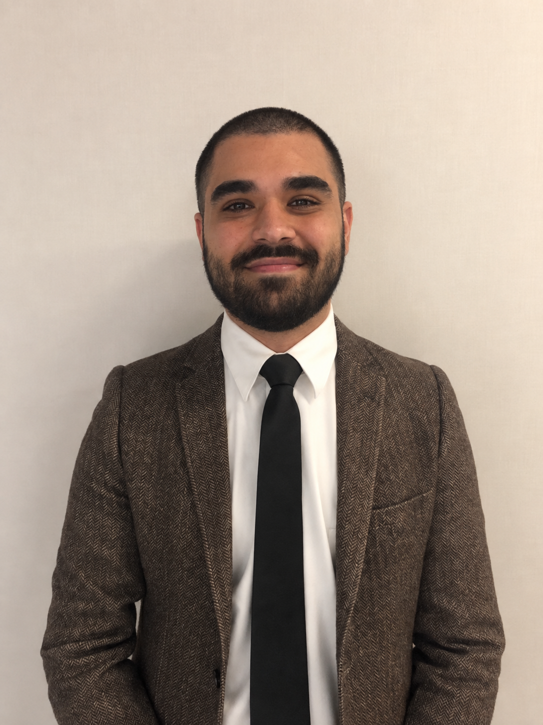 A man with a beard and short hair wearing a white shirt, black tie, and brown blazer standing against a plain wall.