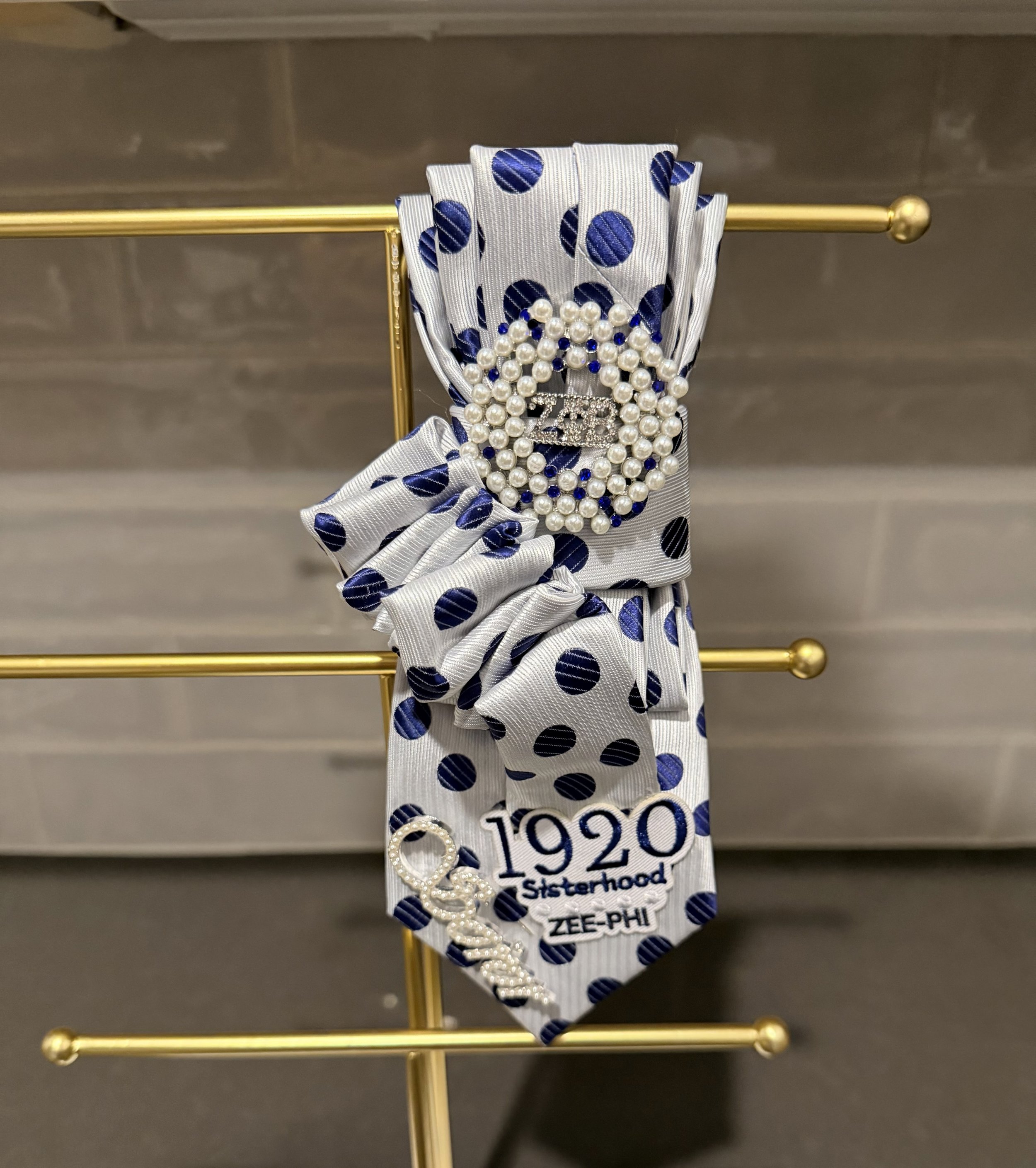 Decorative ribbon with blue polka dots, pearl necklace, and a badge reading '1920 Sisterhood Zee-Phi'.