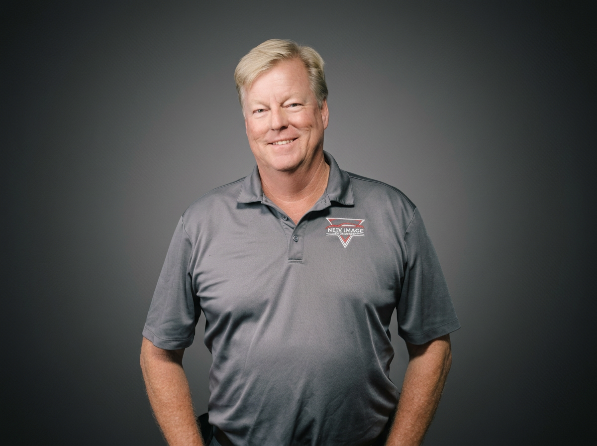 A middle-aged man with blonde hair, smiling, wearing a gray polo shirt with a baseball logo on the left chest area, standing against a dark gray background.