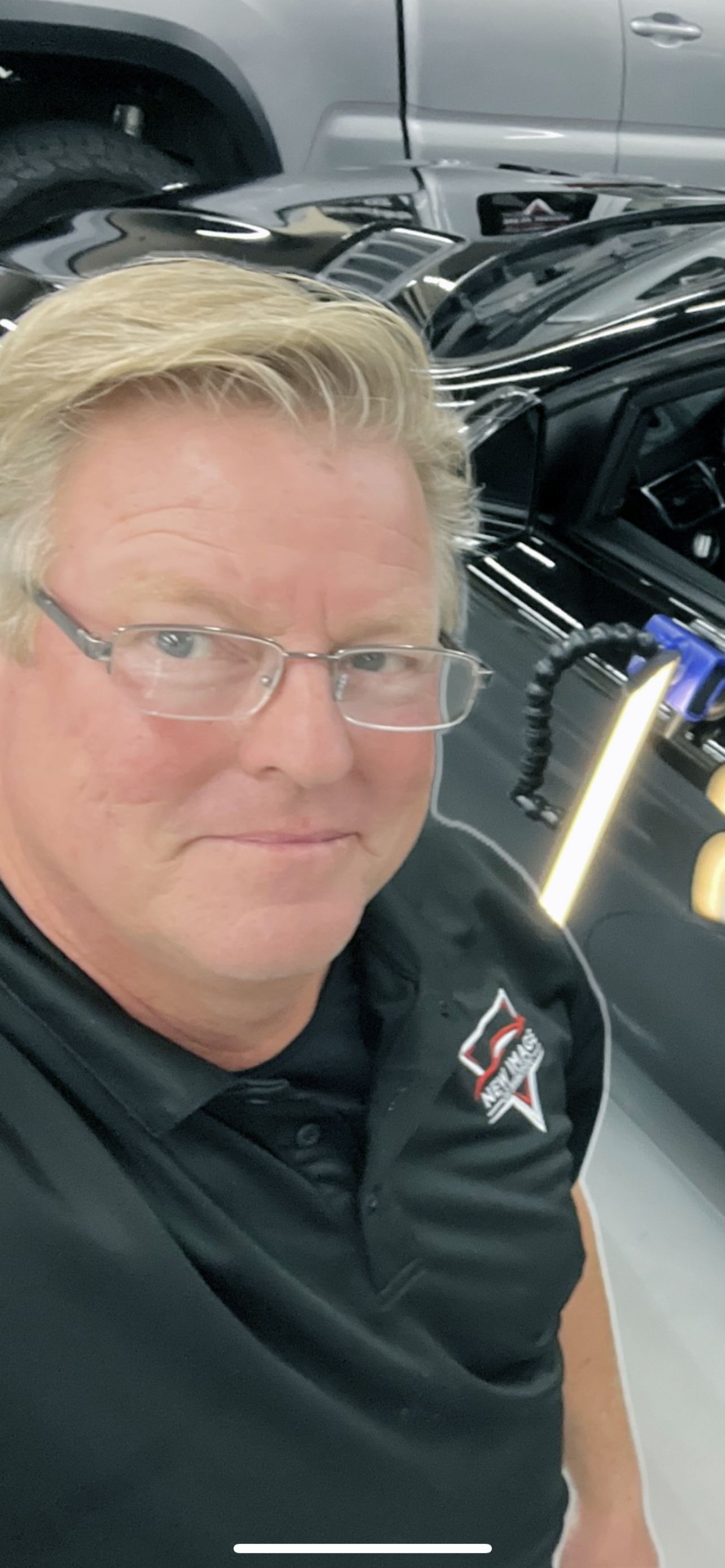 A man with blond hair and glasses taking a selfie performing automotive paintless dent repair in a garage with a black car and another vehicle in the background.