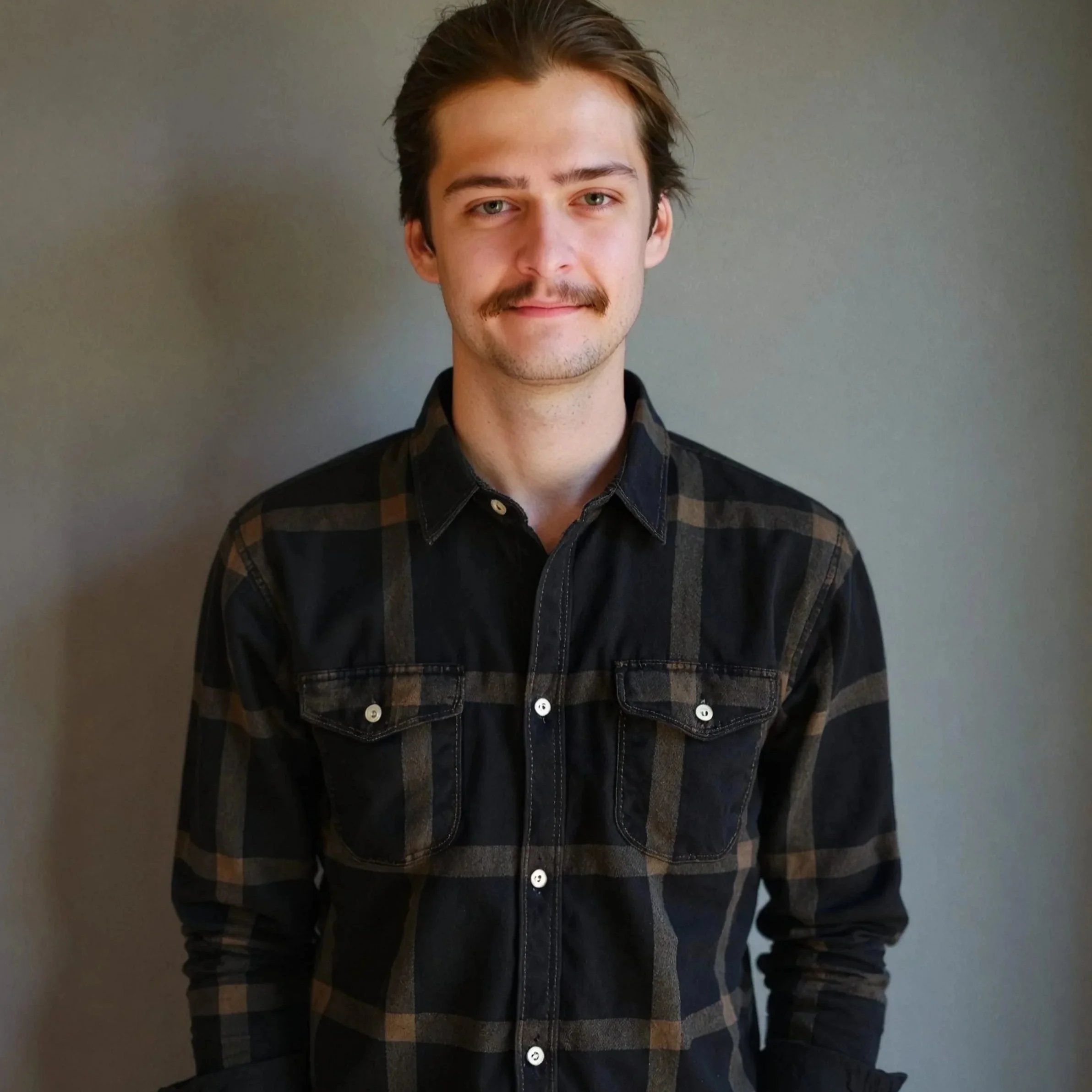 Portrait of Ryan McLendon, wheel repair specialist, a young man with light skin, medium-length brown hair, green eyes, and a mustache, wearing a black and brown plaid shirt, standing against a plain gray wall.