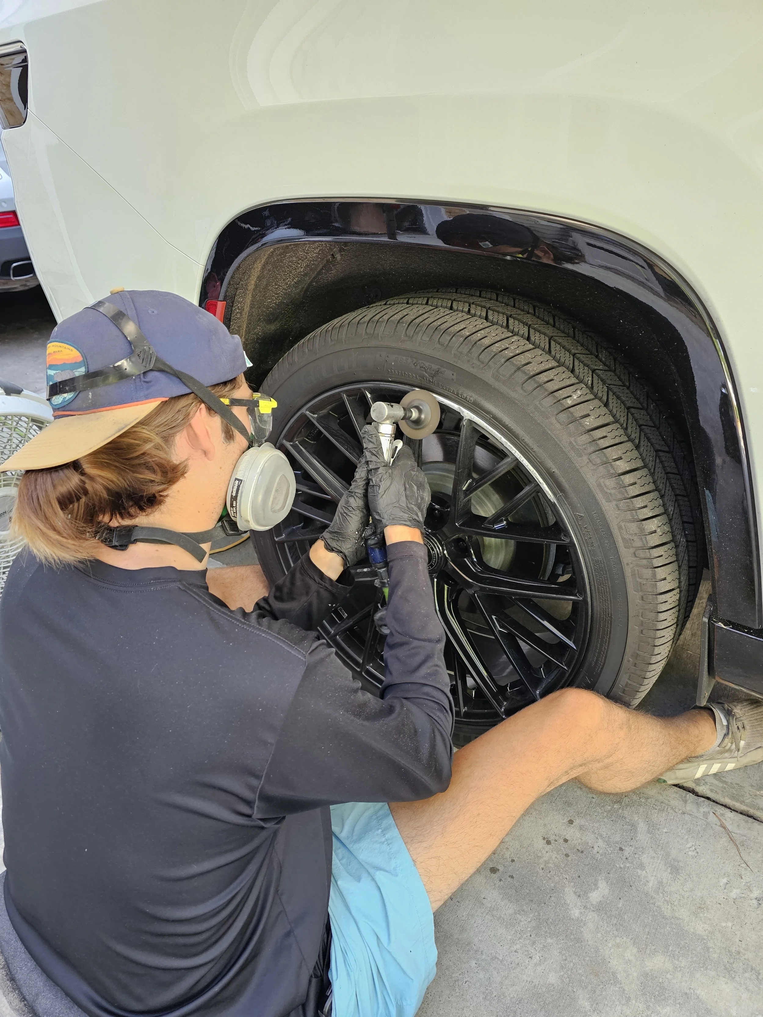A person kneeling on the ground refinishing a car alloy wheel, wearing a wide-brimmed hat, safety glasses, a face mask, and black gloves, using a tool on the wheel of a cream-colored vehicle.