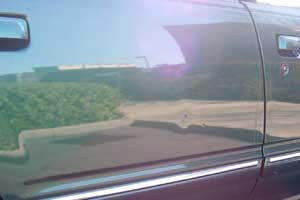 Close-up of ding in a car door with reflections on its surface, showing a grassy area and parking lot.