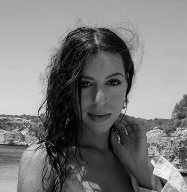 A young woman with wavy dark hair posing outdoors near water and rocks.