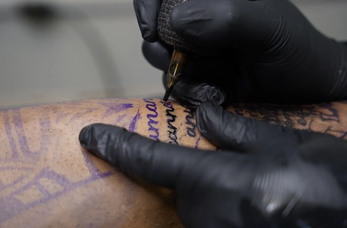A person wearing black gloves is tattooing a design on someone's skin with a tattoo machine, creating black and purple inked lines and letters.