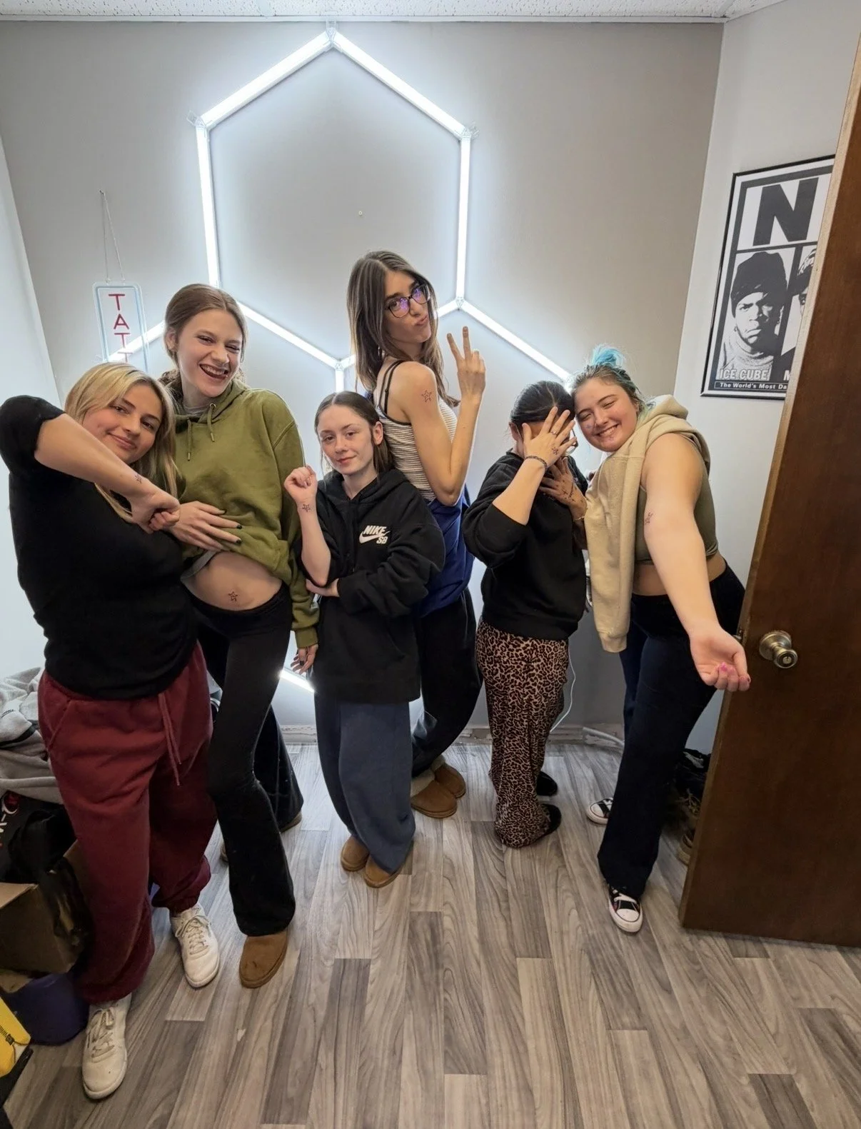 Group of seven young women posing for a photo inside a room with wood flooring and a hexagon-shaped neon light behind them. The women are making showing off their matching tattoos and smiling.