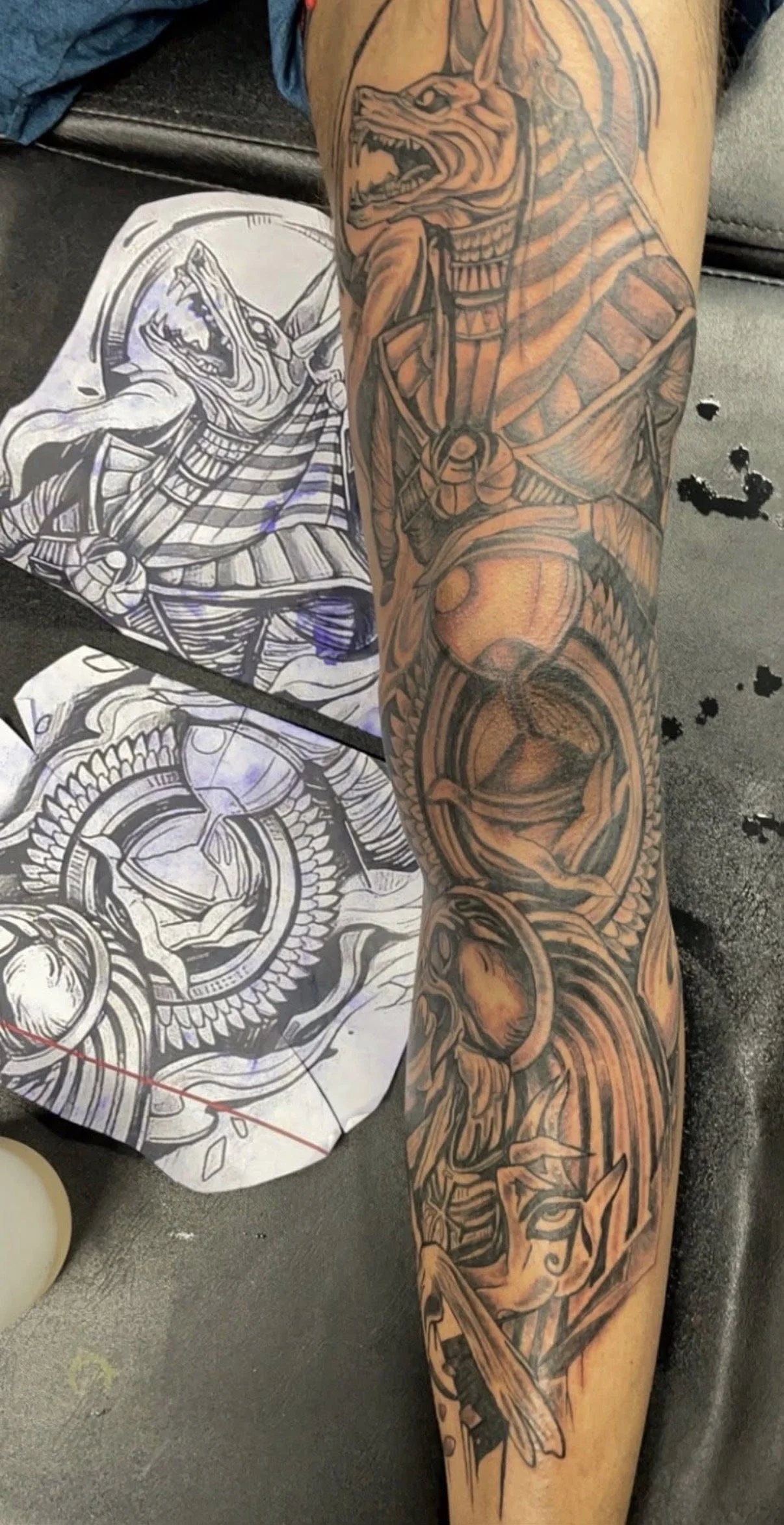 A person's arm with a tattoo of a detailed Egyptian-themed design, including a pharaoh's mask, a scarab beetle, and other Egyptian symbols. Tattoo sketches are seen on paper nearby.