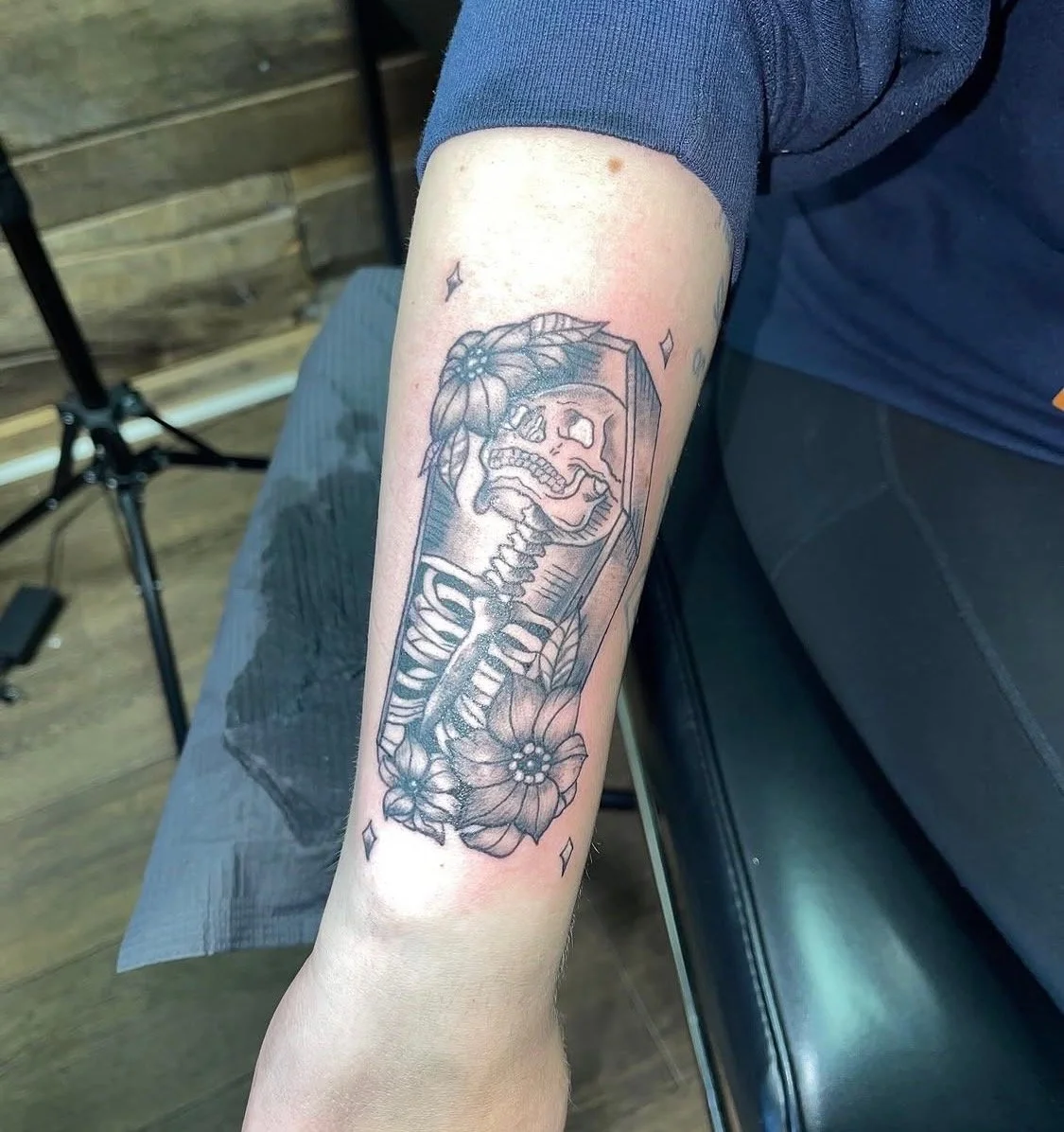 Black tattoo of a human skeleton in a coffin surrounded by flowers on a person's forearm.