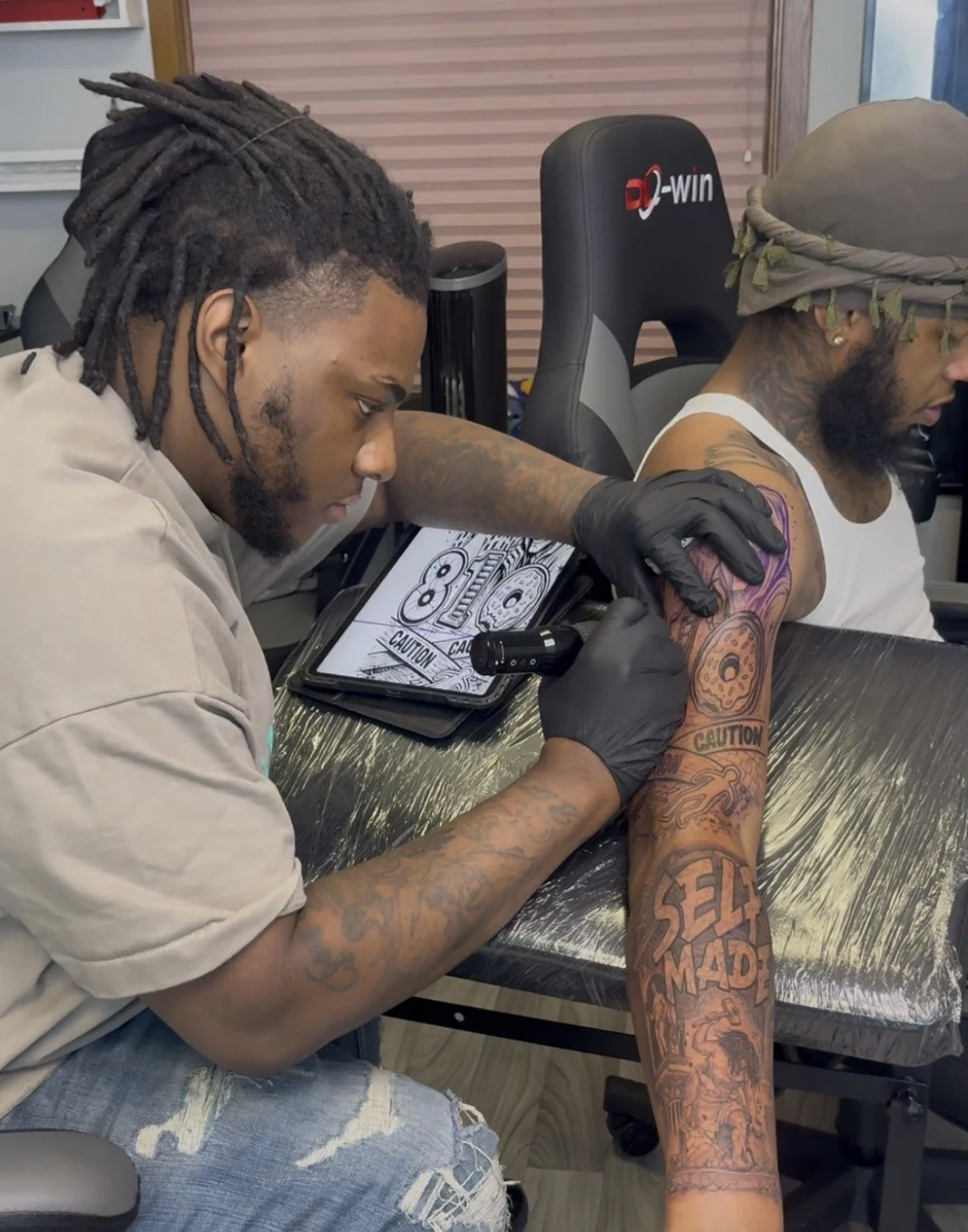 A tattoo artist is tattooing a colorful design on a man's arm while sitting in a chair. The artist is focused, wearing black gloves and a beige shirt. The man has a bandana on his head, sunglasses, and multiple tattoos on his arm and chest.