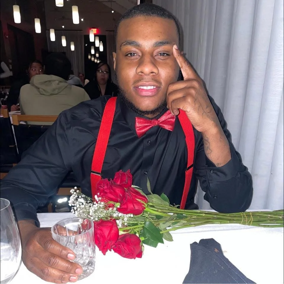 A man with short hair and a beard is sitting at a restaurant table, wearing a black shirt, red suspenders, and a matching red bow tie. He is holding a bouquet of red roses and a glass with a drink. There are other people in the background and the table has white tablecloths and glasses.