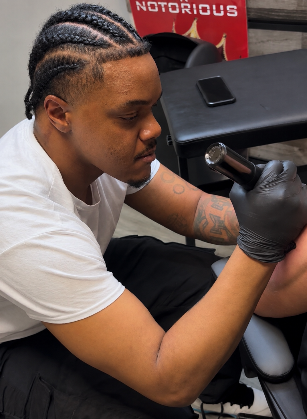 A man with braided hair and tattoos on his arm wearing a white t-shirt and black gloves, sitting at a tattoo station, holding a tattoo machine. There is a black table with a phone on it, and a red poster in the background that reads 'NOTORIOUS'.