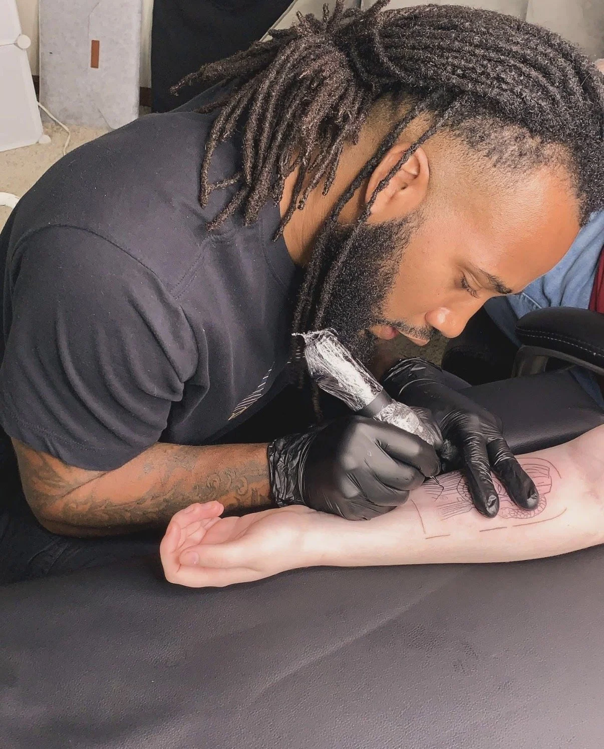 A tattoo artist with dreadlocks and tattoos on his arm is tattooing a design on a person's forearm. The artist is wearing black gloves and is using a tattoo machine. The person receiving the tattoo has light skin.