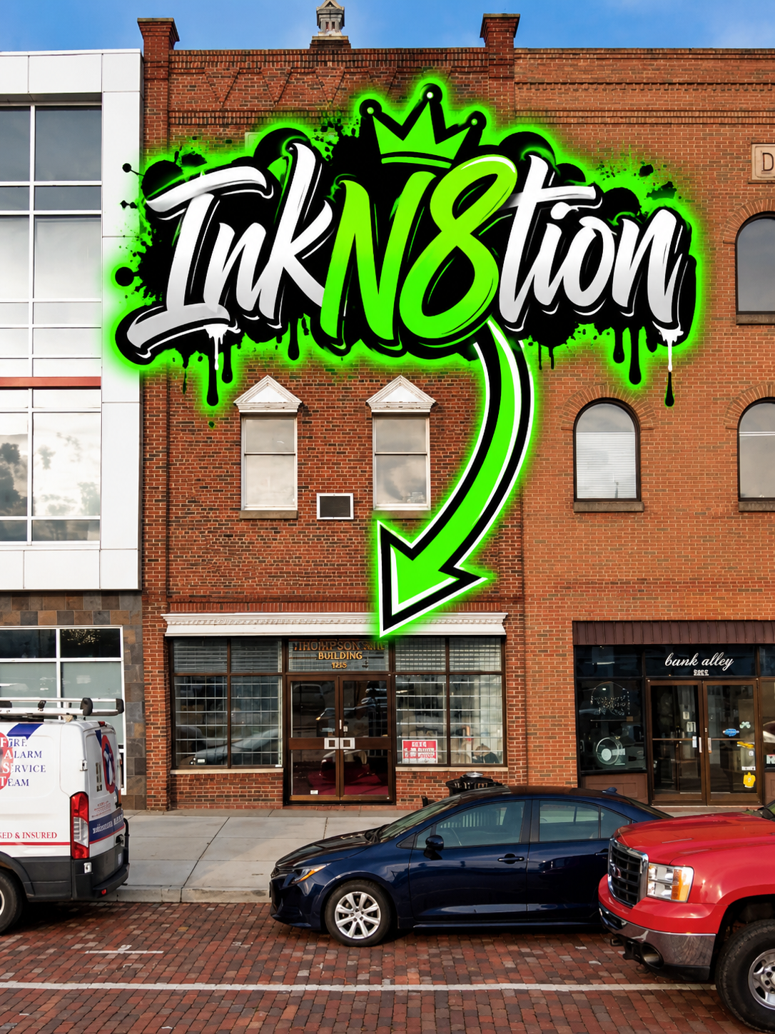 Graffiti-style sign on a brick building reads 'InkN8tion' with a green arrow pointing to the storefront below. The building has large glass windows and neighboring storefronts, parked cars on the street.