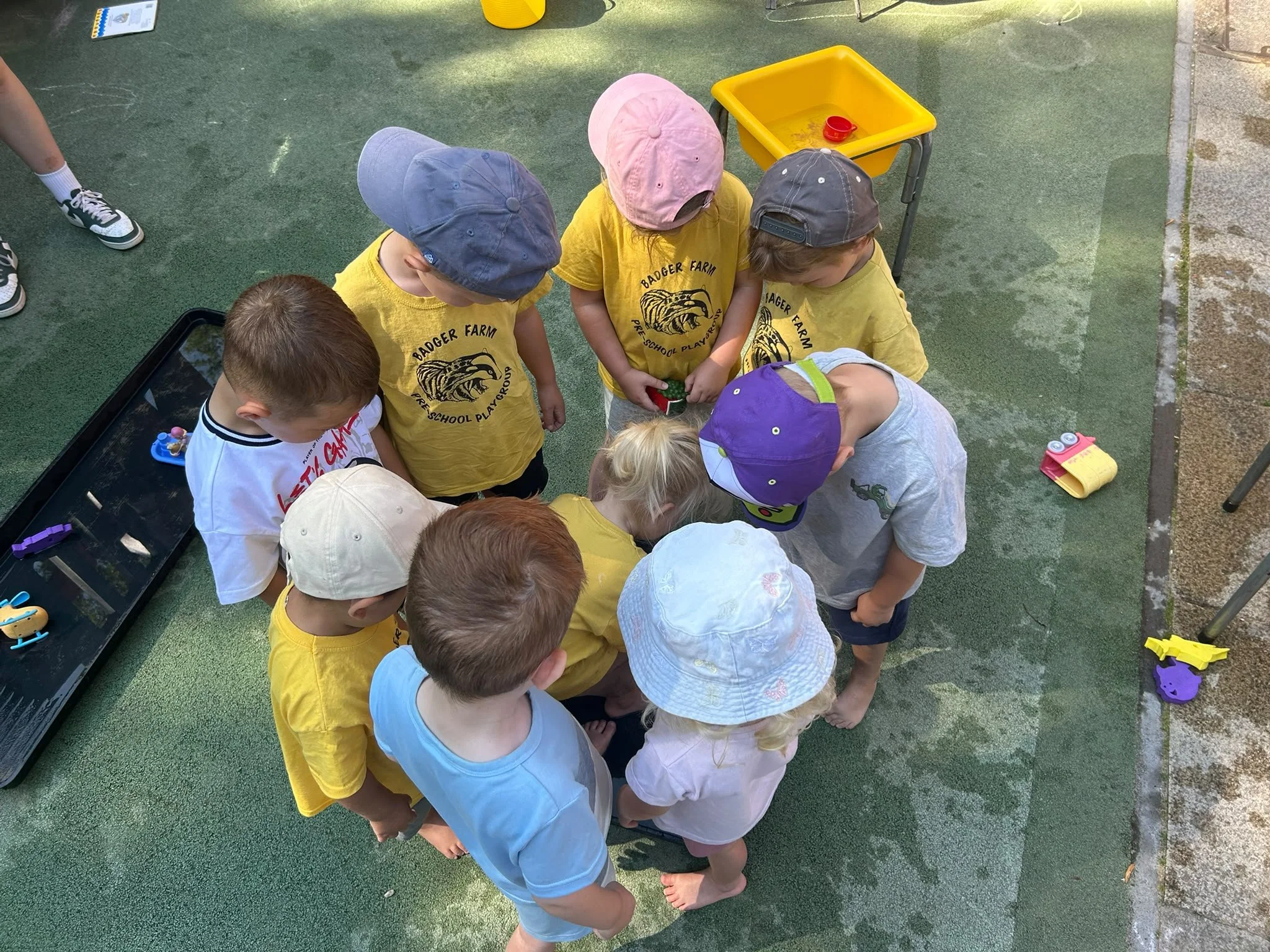 Children in yellow shirts and colorful hats gathered around, looking at something on the ground outside. A yellow container and some toys are nearby.