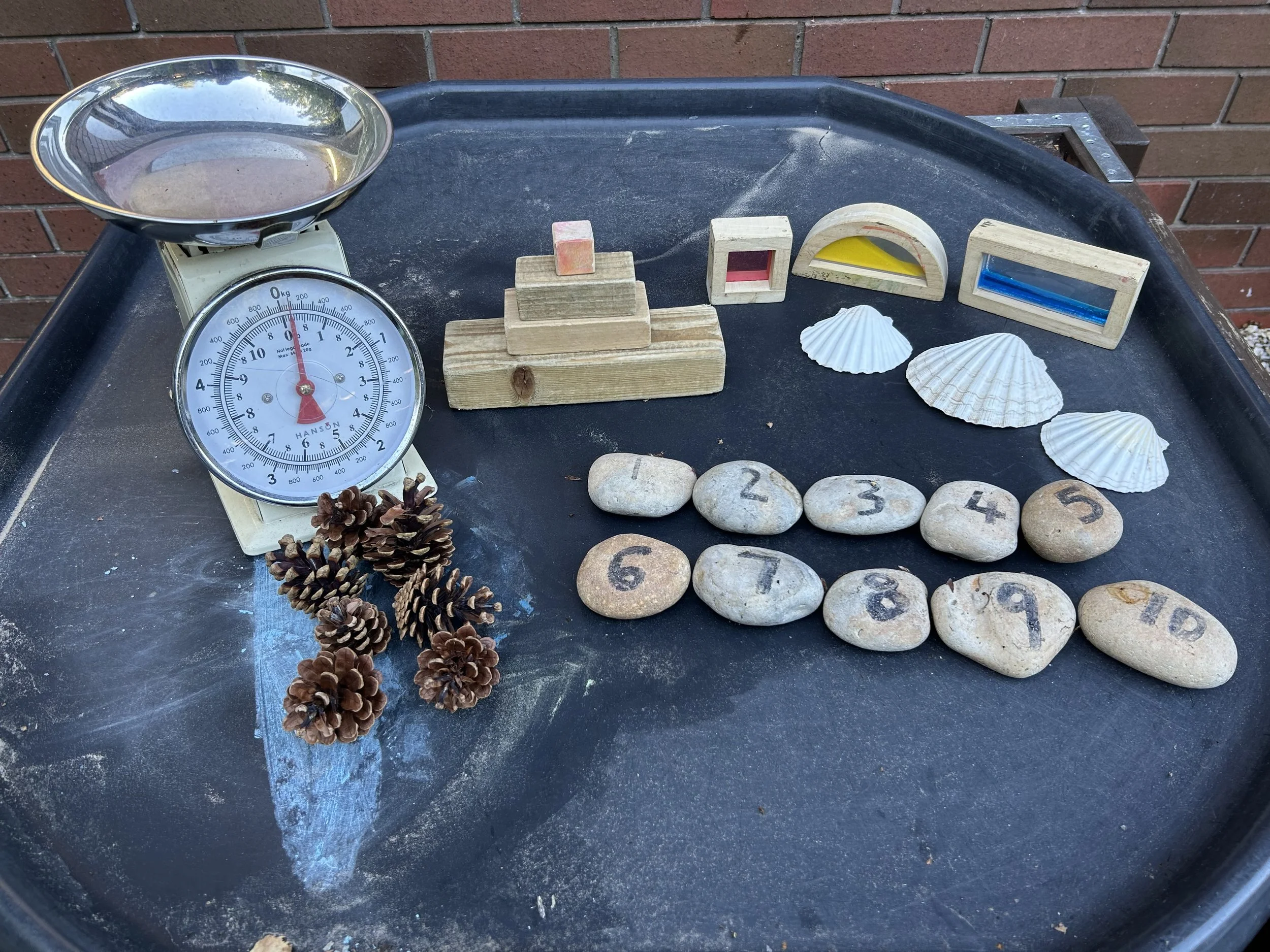 A black tray with a vintage balance scale, decorative pinecones, seashells, numbered stones, and various wooden geometric shapes.