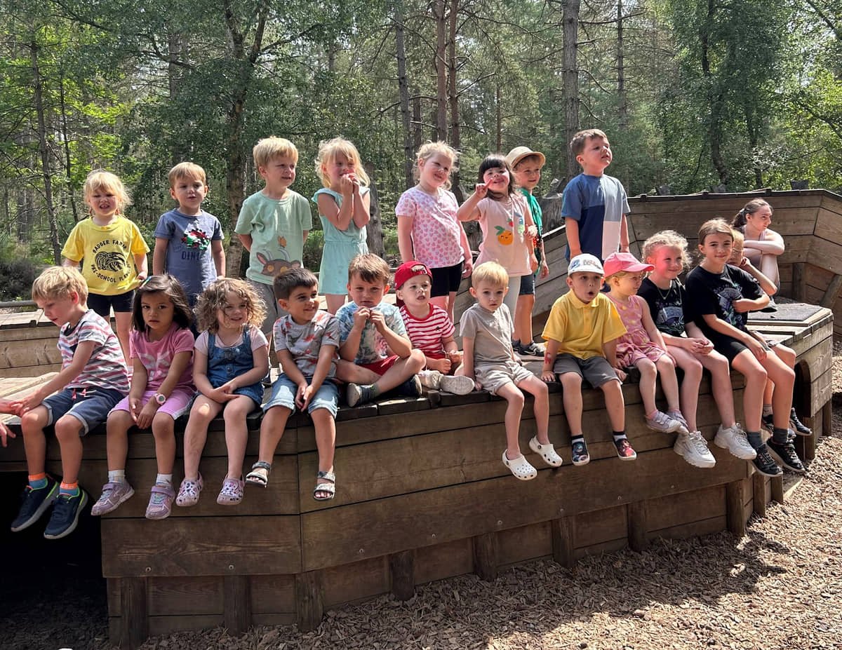 Group of young children sitting and standing on a wooden structure outdoors in a forest, smiling and looking at the camera.