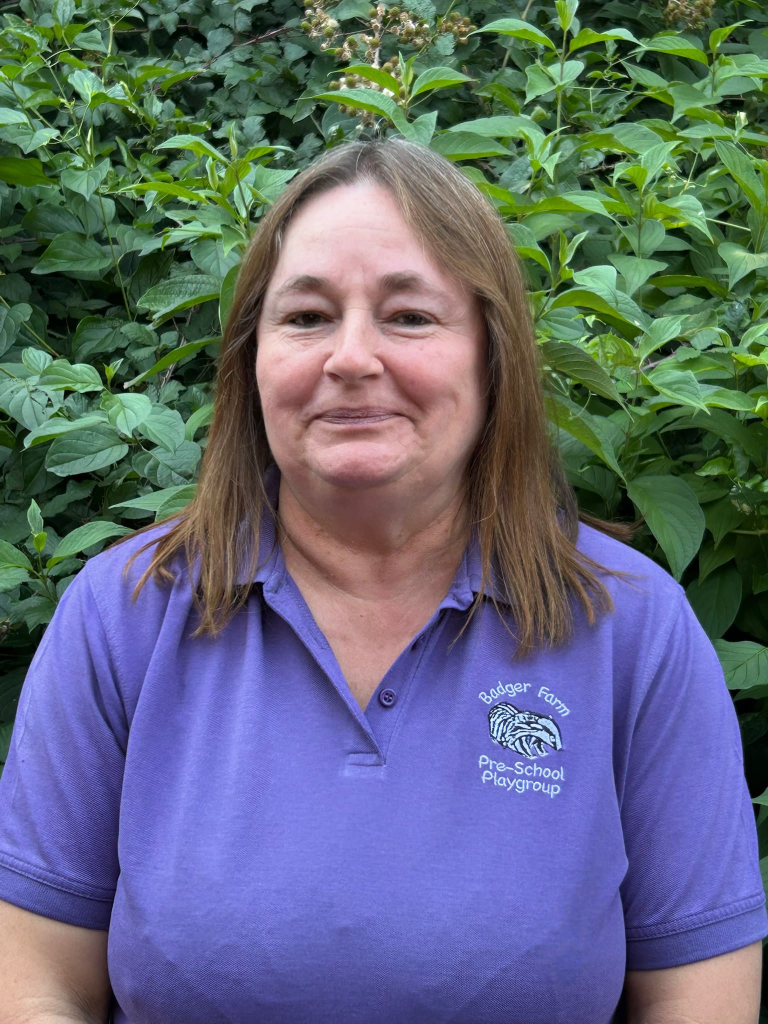 Close-up of a woman in a purple polo shirt with 'Badger Farm Pre-School Playgroup' logo, standing in front of green leafy bushes, smiling slightly.