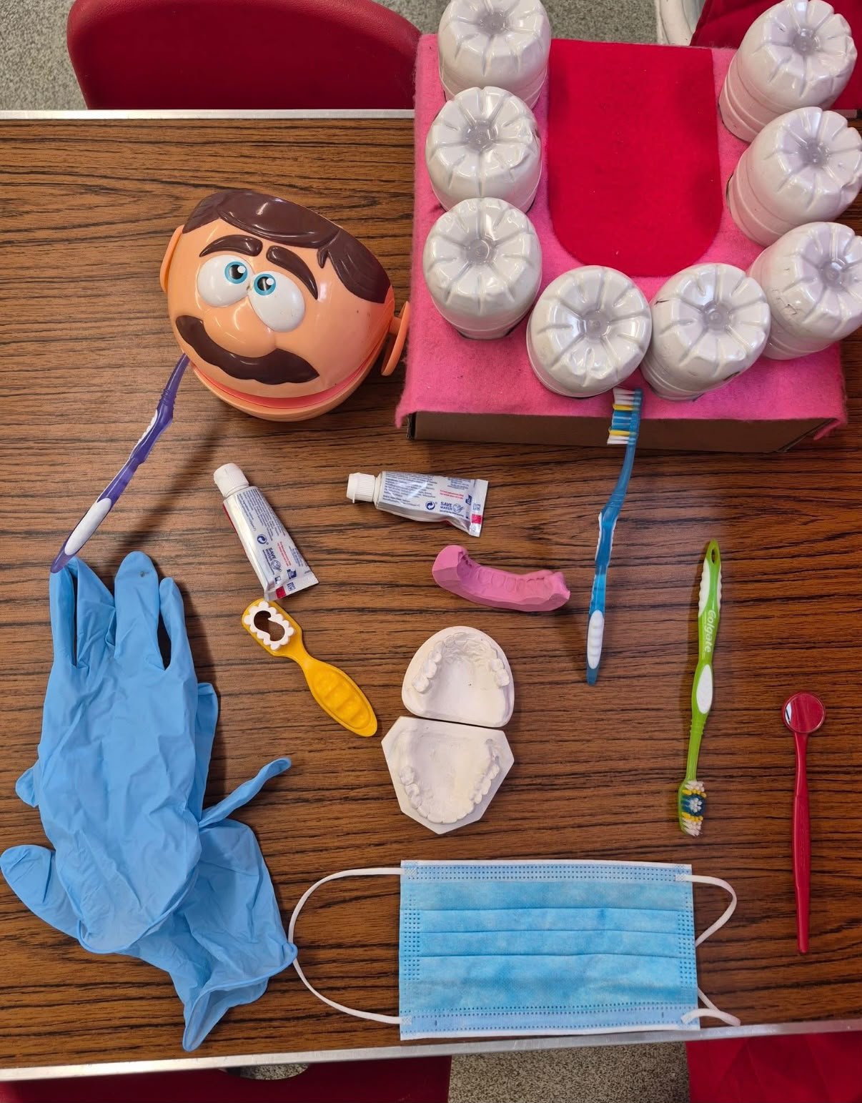 A table filled with various dental hygiene and dental practice items, including a Mr. Potato Head toy with a toothbrush in its mouth, white dental molds, a disposable face mask, toothbrushes, toothpaste tubes, gloves, and other dental tools.