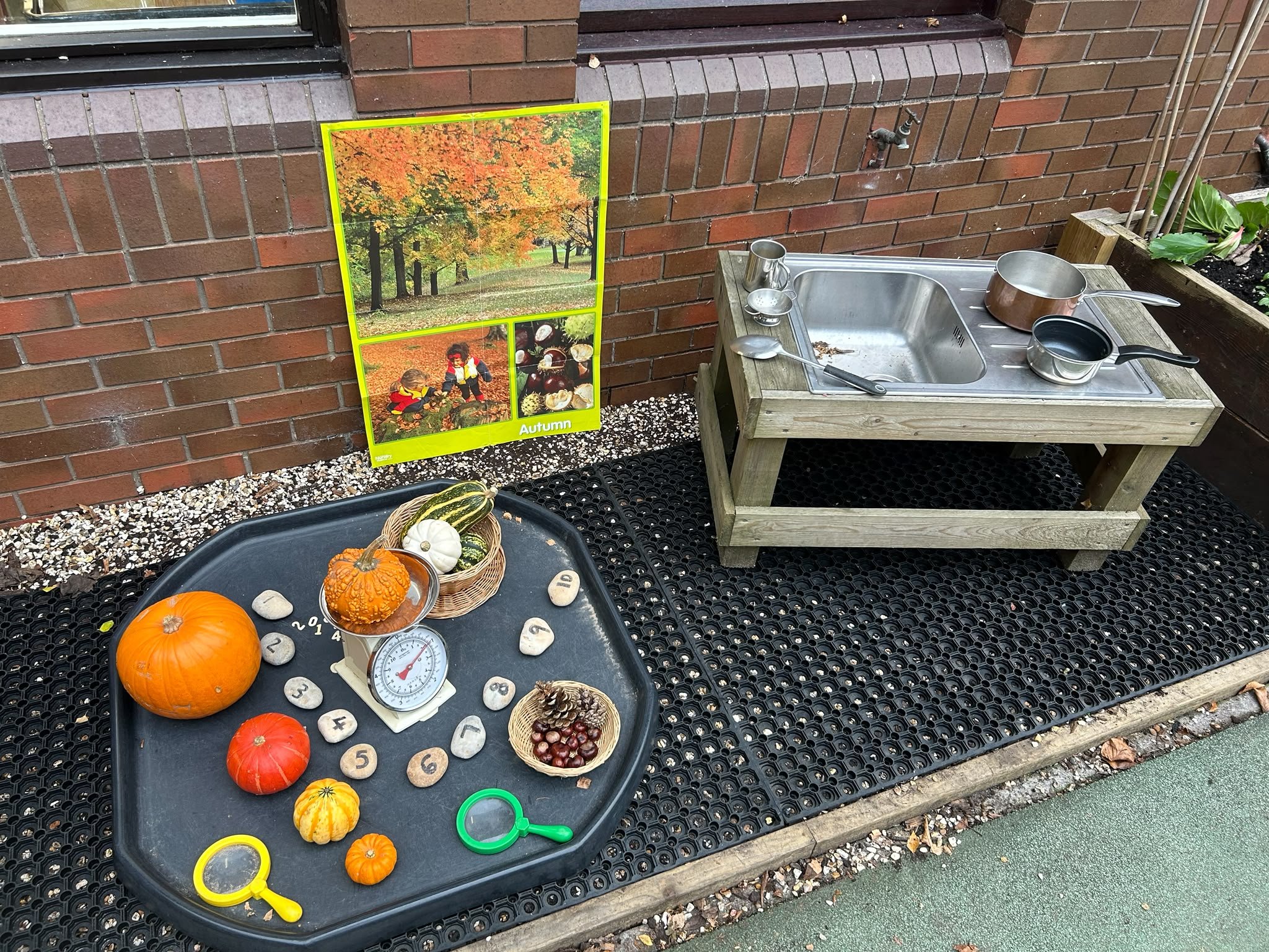 A small outdoor sink with cleaning supplies, next to a potted plant, and a colorful poster of autumn trees with children playing in fallen leaves. On the black surface below, there is a tray with small pumpkins, a timer, and some scattered stones.