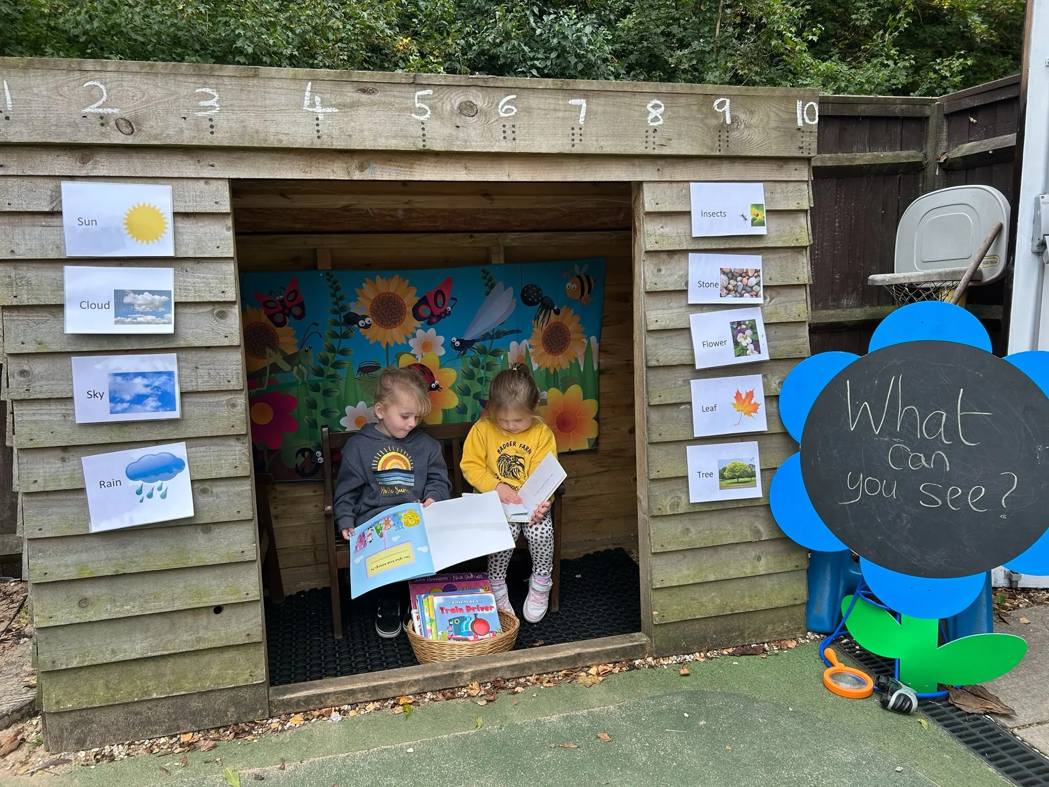 Children reading books inside a wooden playhouse with educational signs displaying weather-related words and images, outside in a park or playground area.