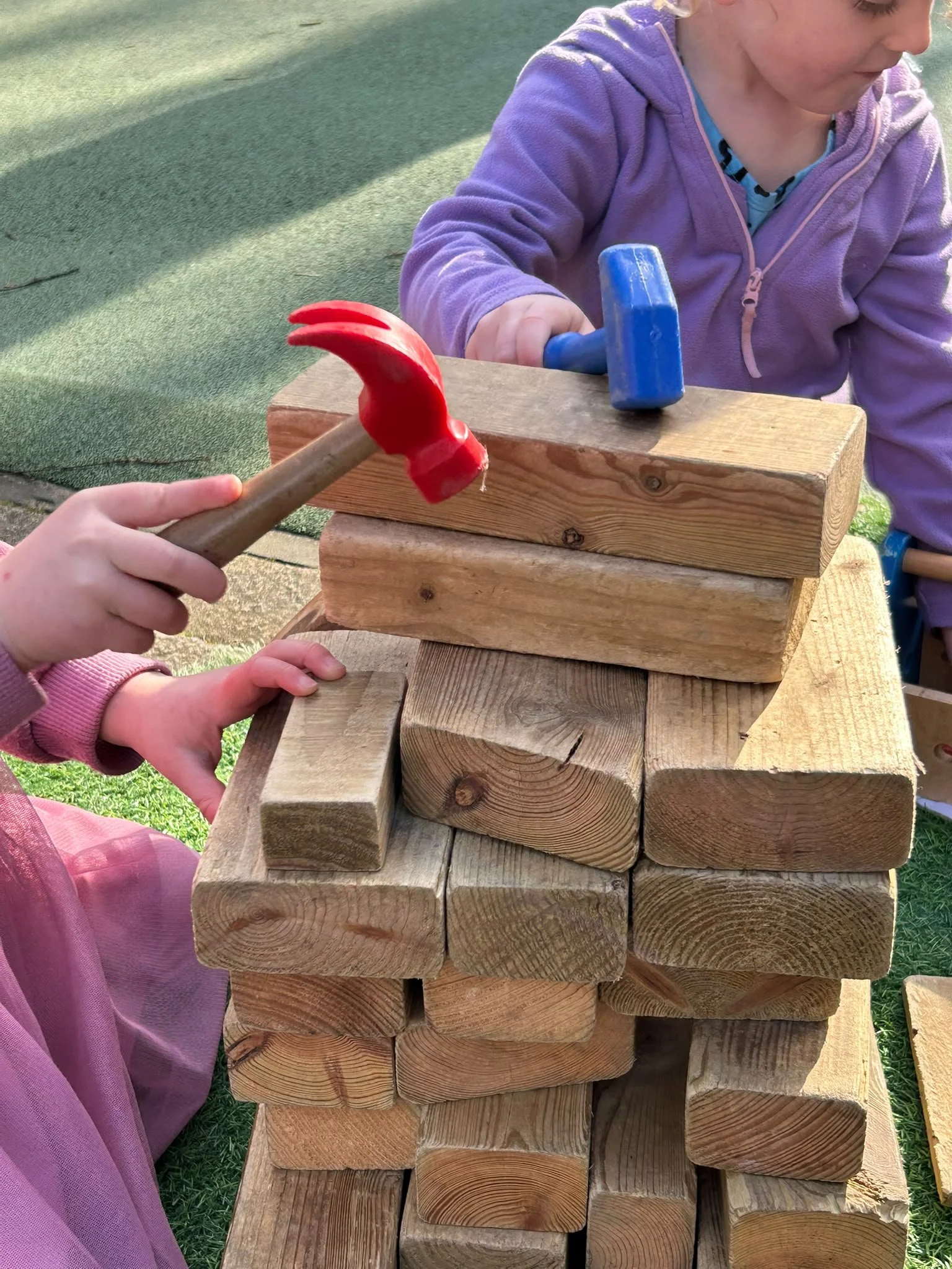 Children stacking wooden blocks outdoors, with one child holding a toy hammer, wearing a purple jacket, part of a childcare or play activity.