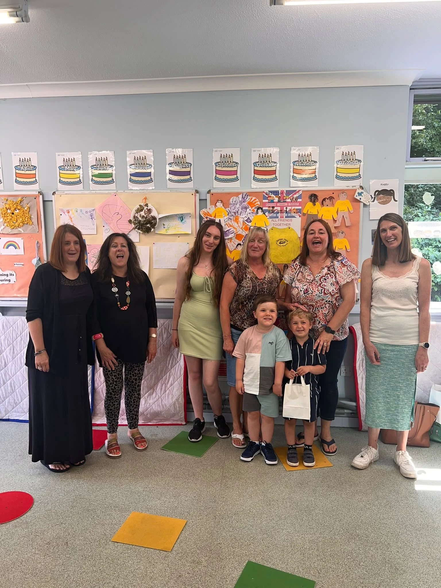 A group of seven women and three children standing in a room with educational decorations and colorful charts on the wall. The women are smiling and the children are smiling, with some standing on colorful floor squares.