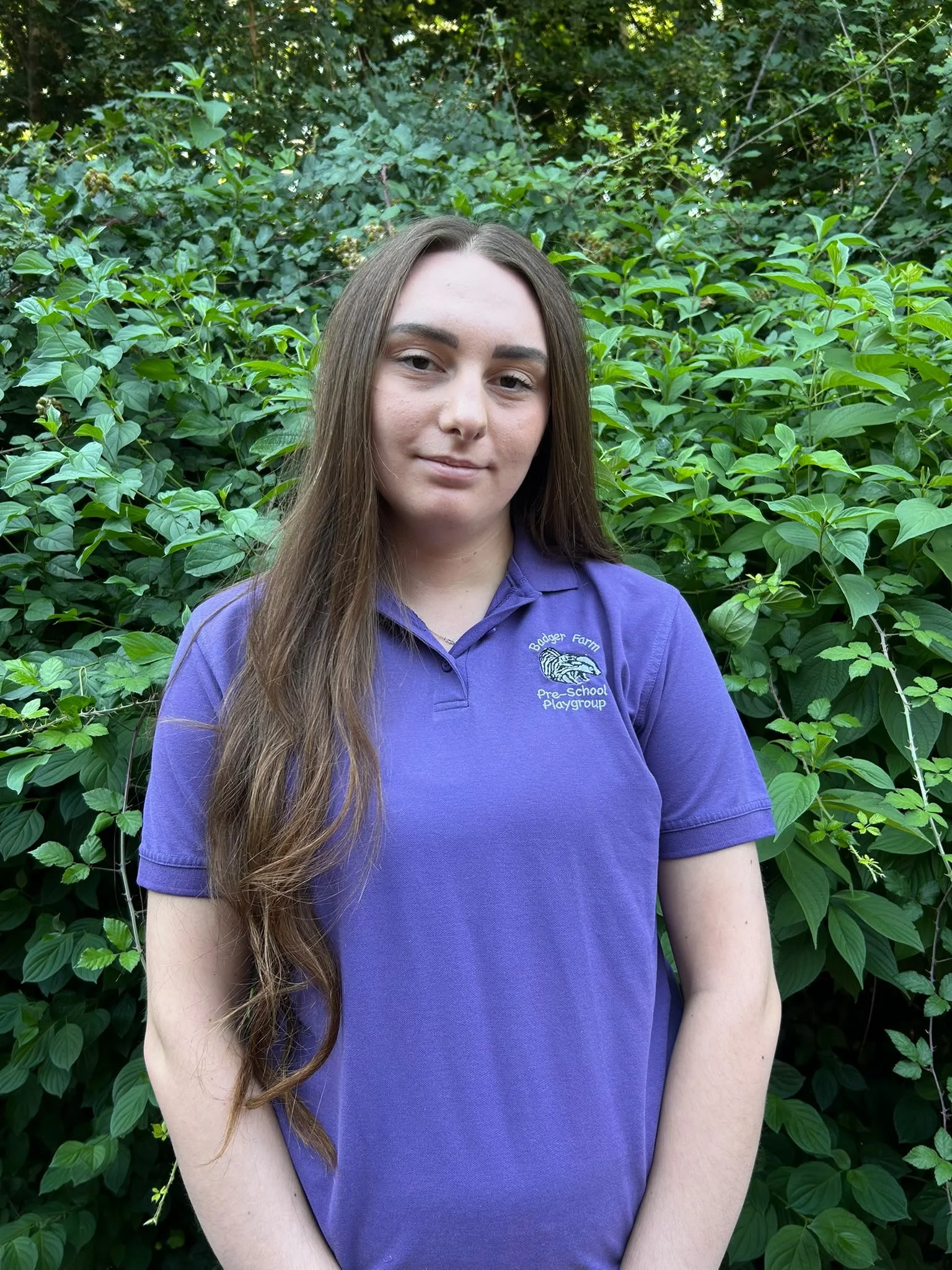 A young woman with long brown hair wearing a purple polo shirt stands in front of lush green foliage.
