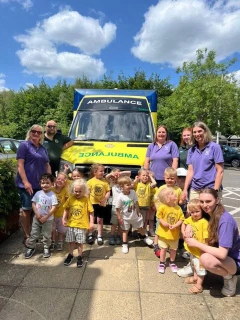 Group of children and adults standing in front of an ambulance, outdoor setting with trees and blue sky.
