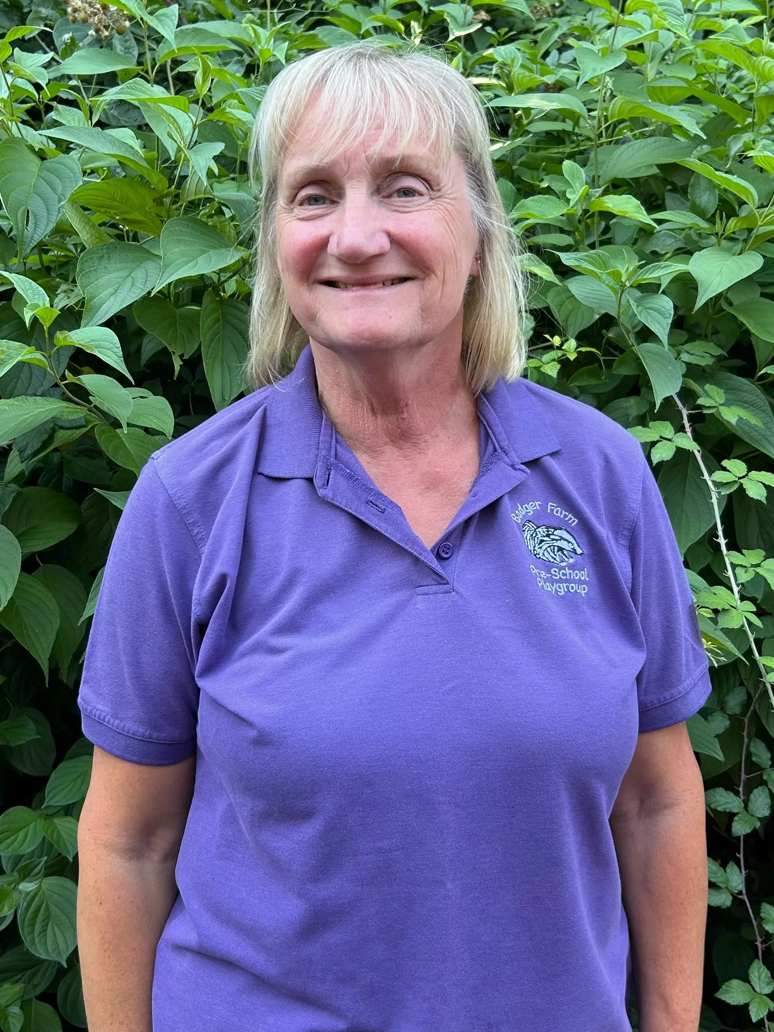 A woman with blonde hair wearing a purple polo shirt standing in front of green foliage.