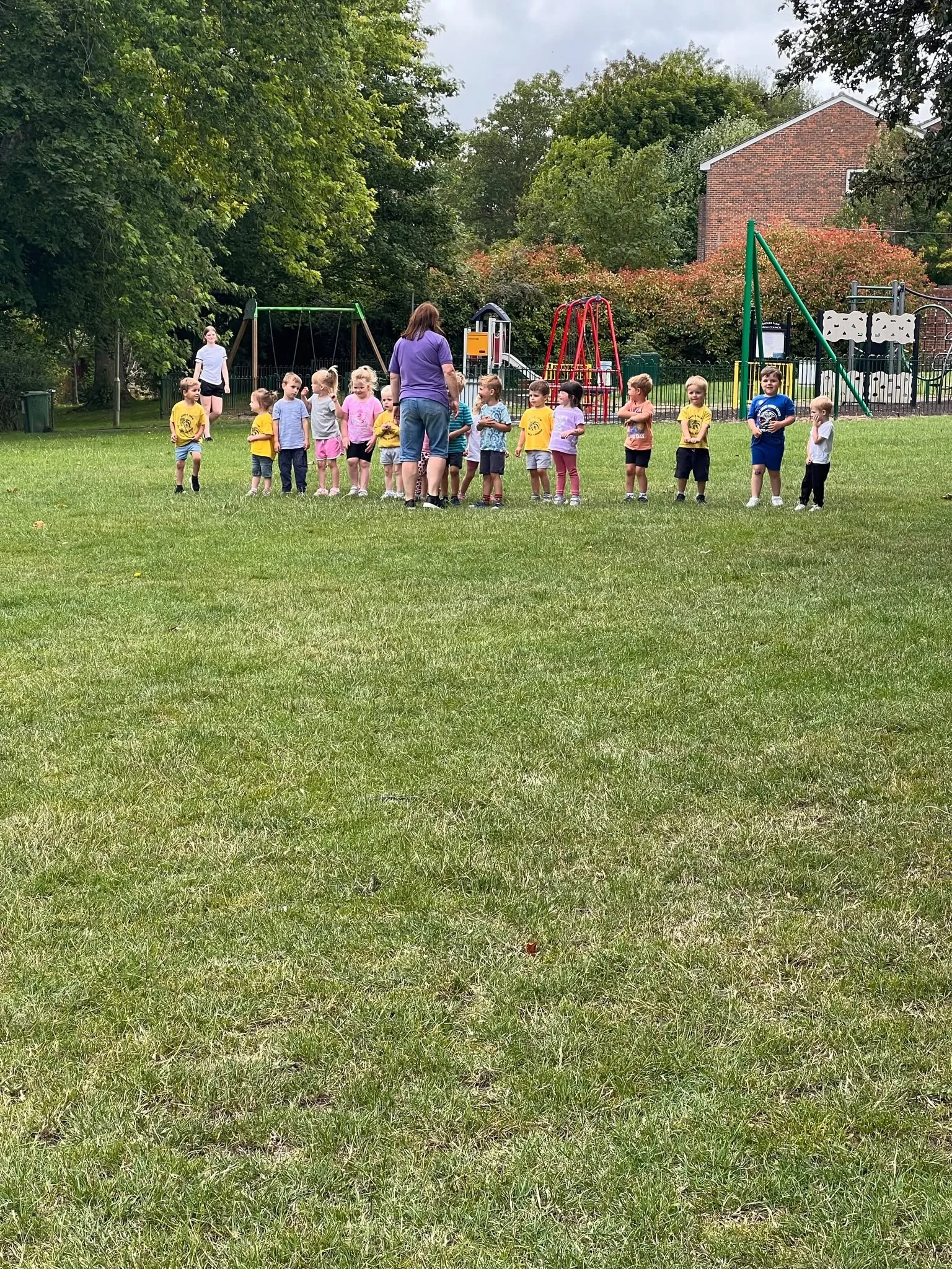 A group of young children are gathered outdoors on a grassy field, accompanied by two adults. The children are standing in a line, some wearing yellow shirts, and seem to be participating in an outdoor activity. In the background, there are playgroun