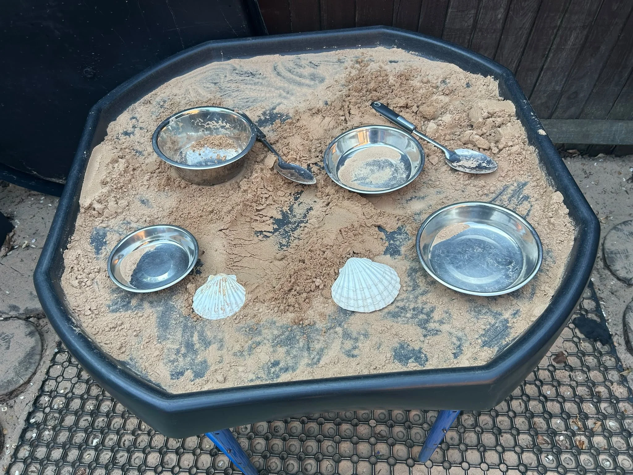 A black metal tub filled with sand, with several small metal bowls and spoons placed on top, some filled with sand. Two large shells are also on the sand.