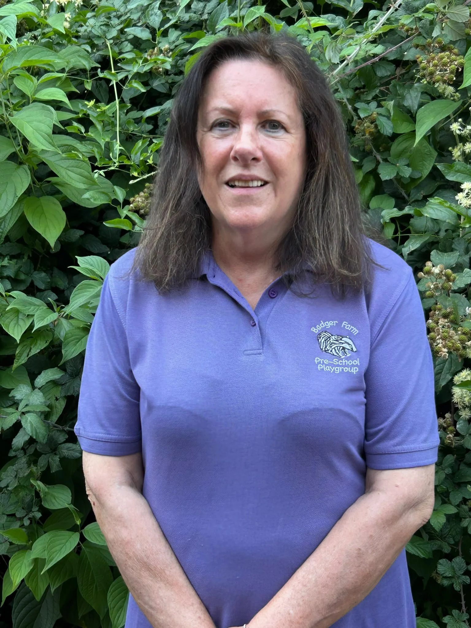 A woman with brown hair wearing a purple polo shirt with an embroidered logo that reads "Badger Farm Pre-School Playgroup". She is standing outdoors in front of green leafy foliage.