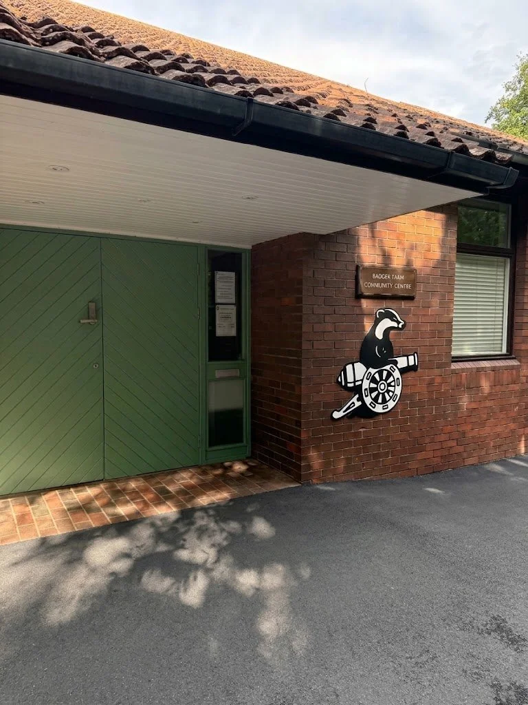 The entrance to the Badger Taan Community Centre, featuring a large green door, brick walls, and a black mascot figure of a badger with a wheel and a rod, mounted next to a sign reading 'Badger Taan Community Centre'.