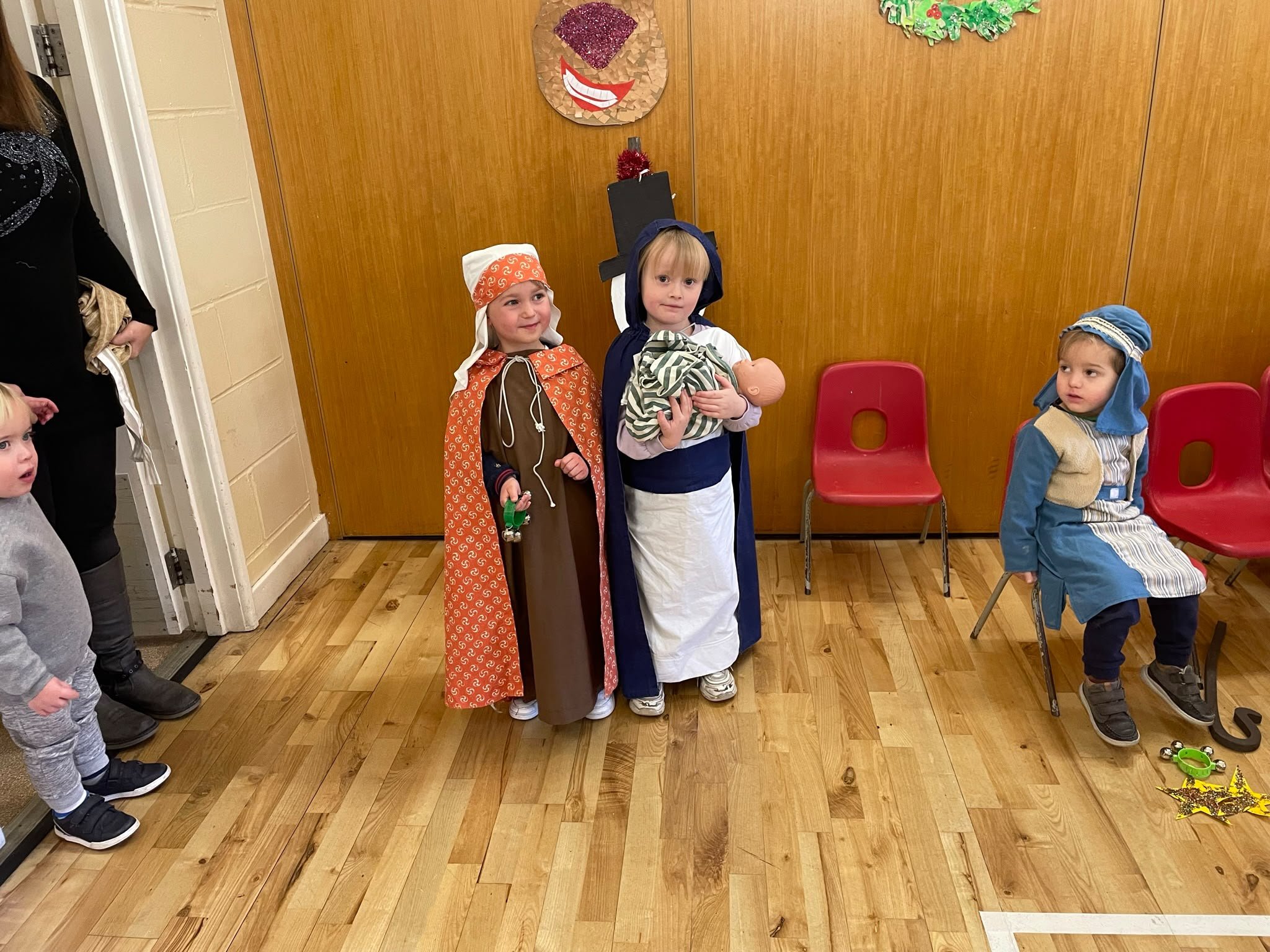 Children dressed in costumes, standing and sitting in a room decorated for the holidays, with a Christmassy wreath and smiling face decoration on the wall behind them.