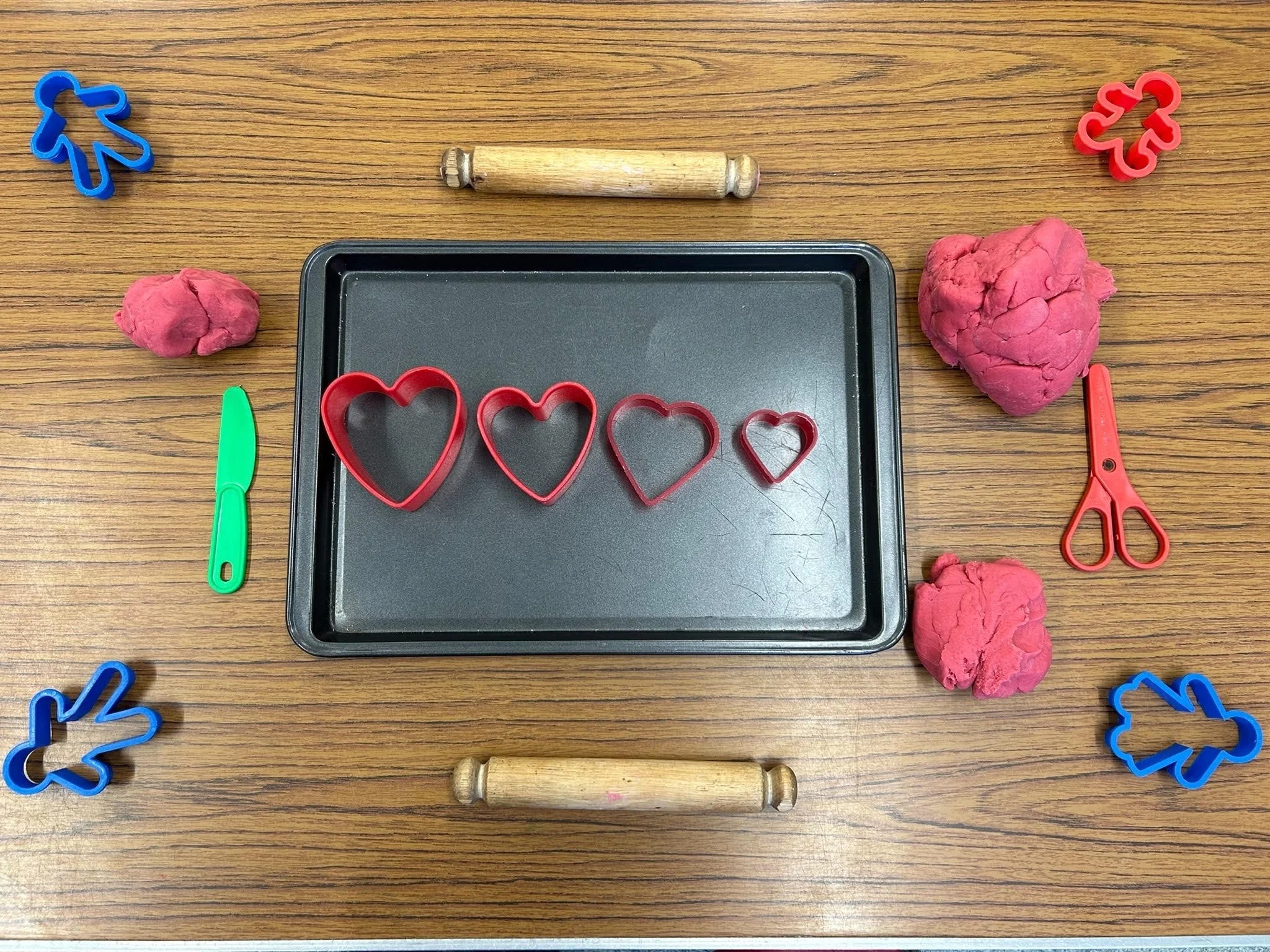 Plastic cookie cutters, two rolling pins, two balls of pink clay, a green plastic knife, and a red pair of scissors are arranged on a wooden table.