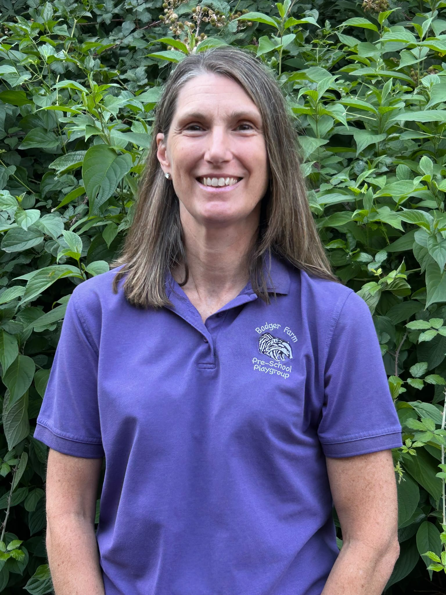 A smiling woman with long brown hair wearing a purple polo shirt standing in front of leafy green plants.