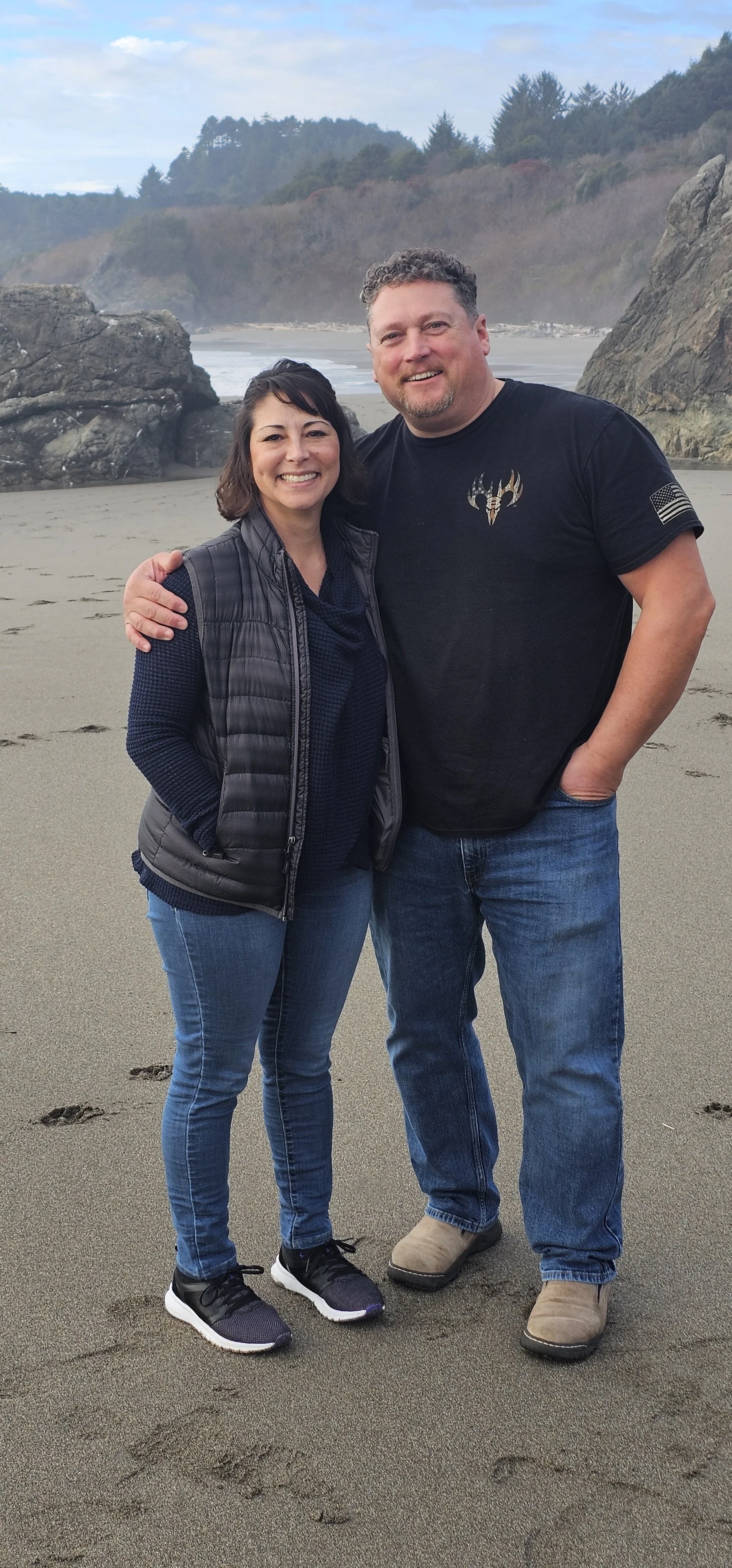 A smiling woman and man standing close together on a sandy beach, with large rocks and trees in the background.
