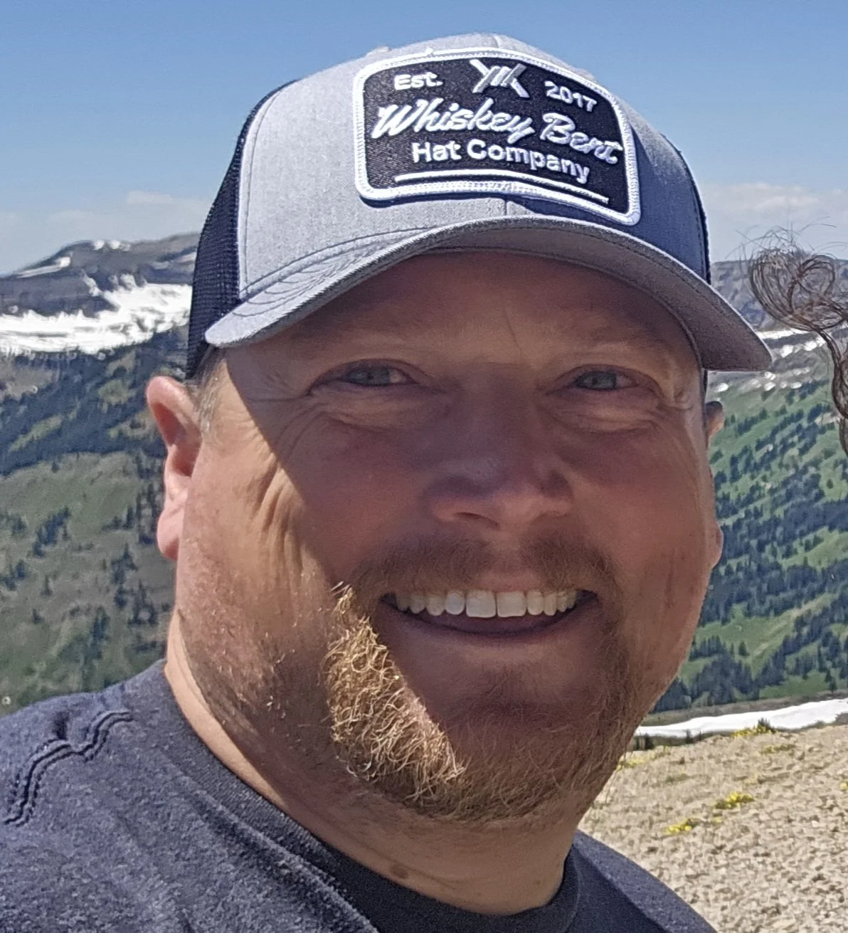 A smiling man with a beard wearing a gray and navy trucker hat with a whiskey barrel company logo, standing outdoors with snow-capped mountains and green hills in the background.