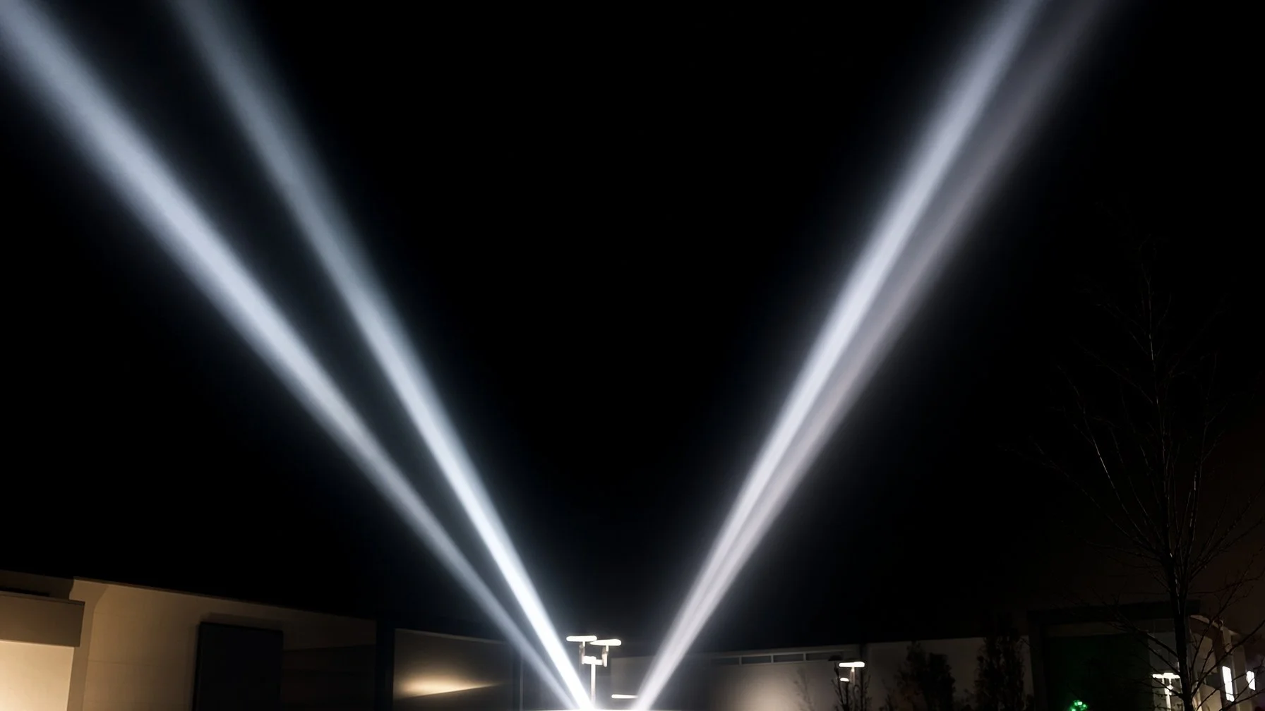 Nighttime outdoor scene with four bright white beams of light crossing at a fountain, illuminating the sky.