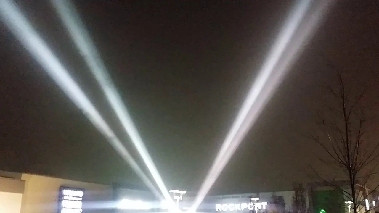 Bright beams of light shine upward into the night sky. Some trees and buildings are visible in the foreground, with a sign that reads "ROCKPORT."