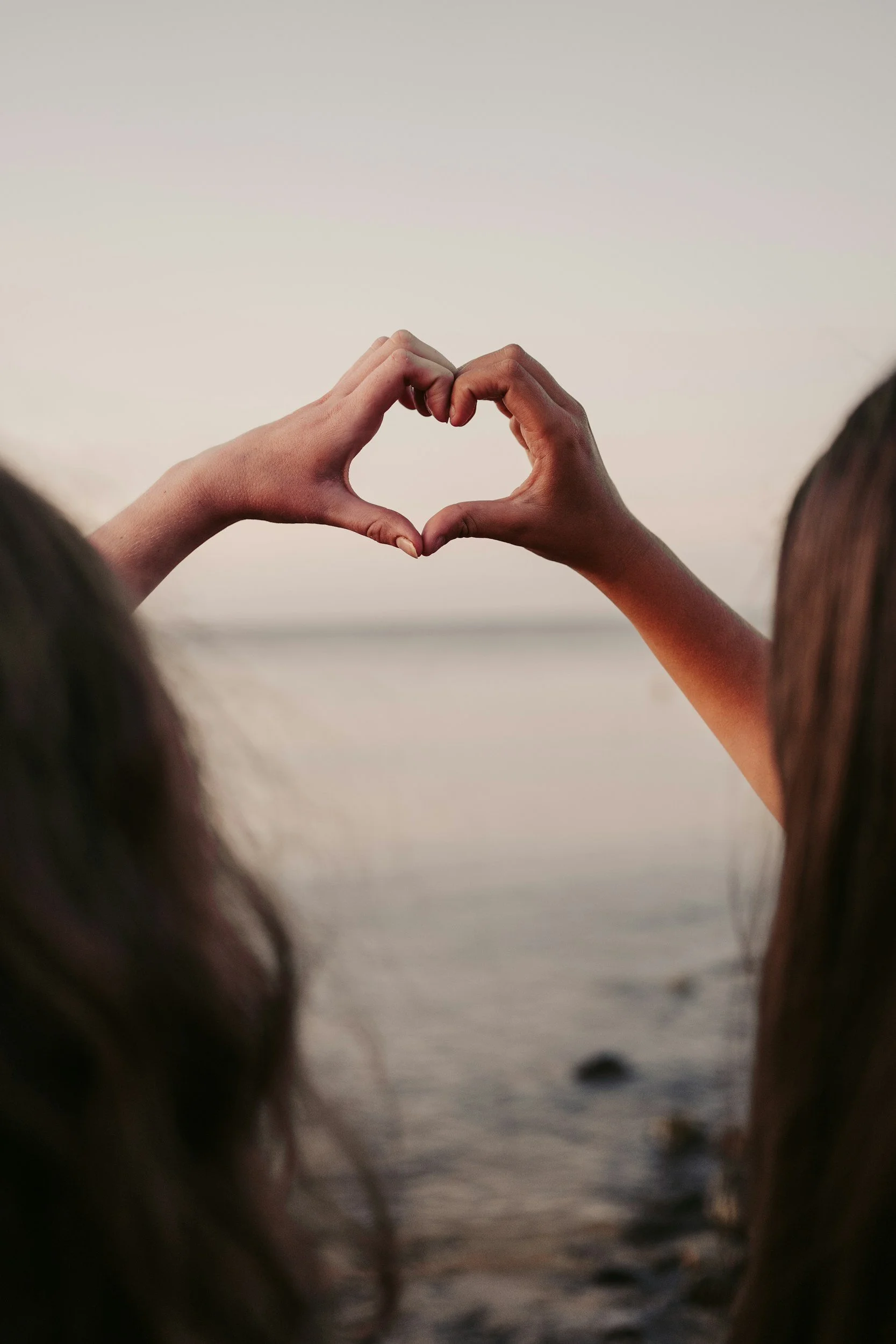 Two hands forming a heart shape against a beach at sunset, with hair visible in the foreground.