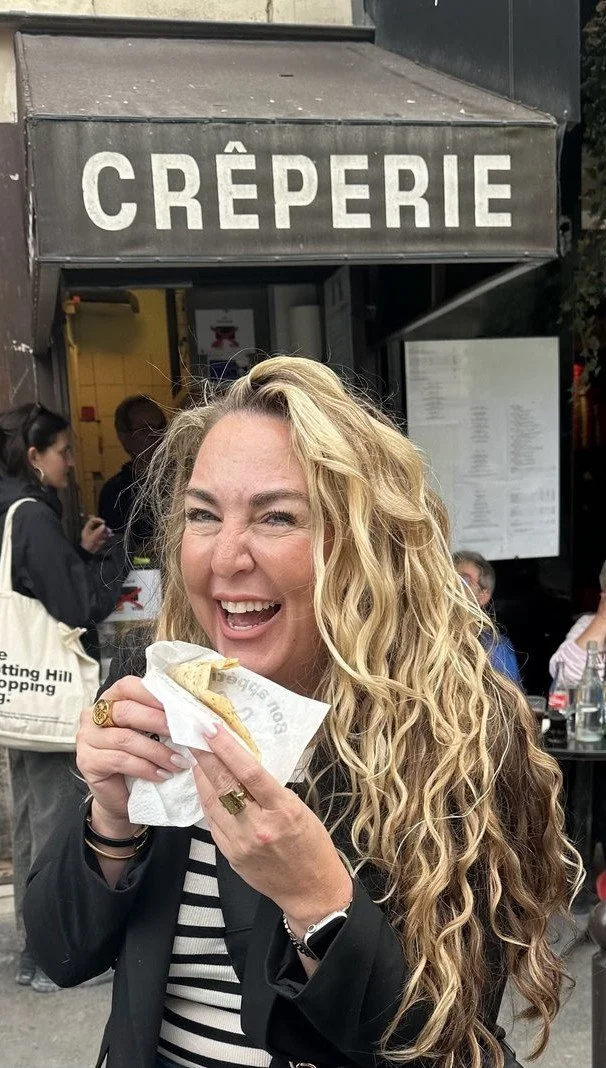 Woman with long curly blonde hair smiling and holding a crepe outside a creperie.