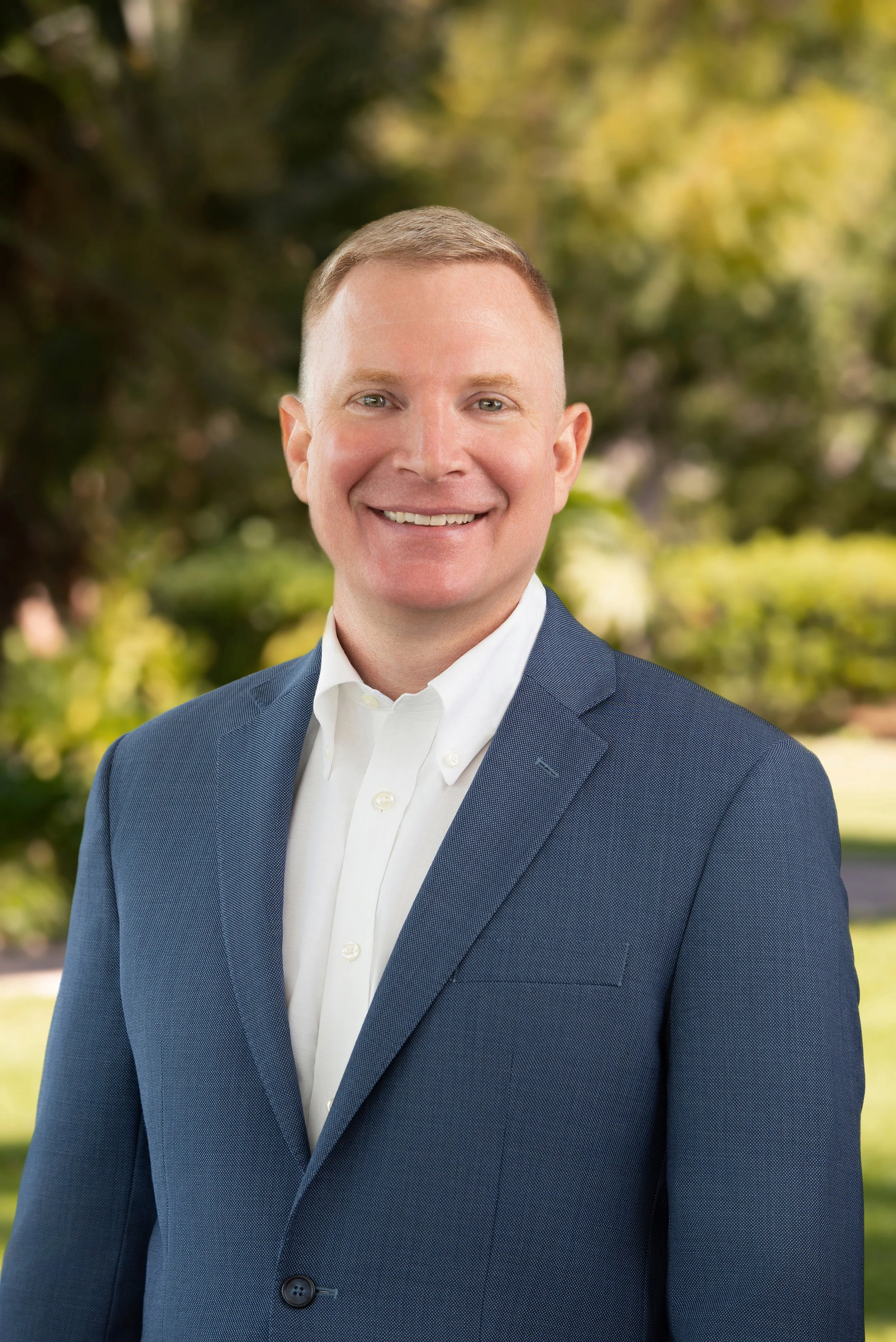 Headshot of a smiling man with short hair, wearing a blue suit jacket and white dress shirt, outdoors with blurred green trees in the background.