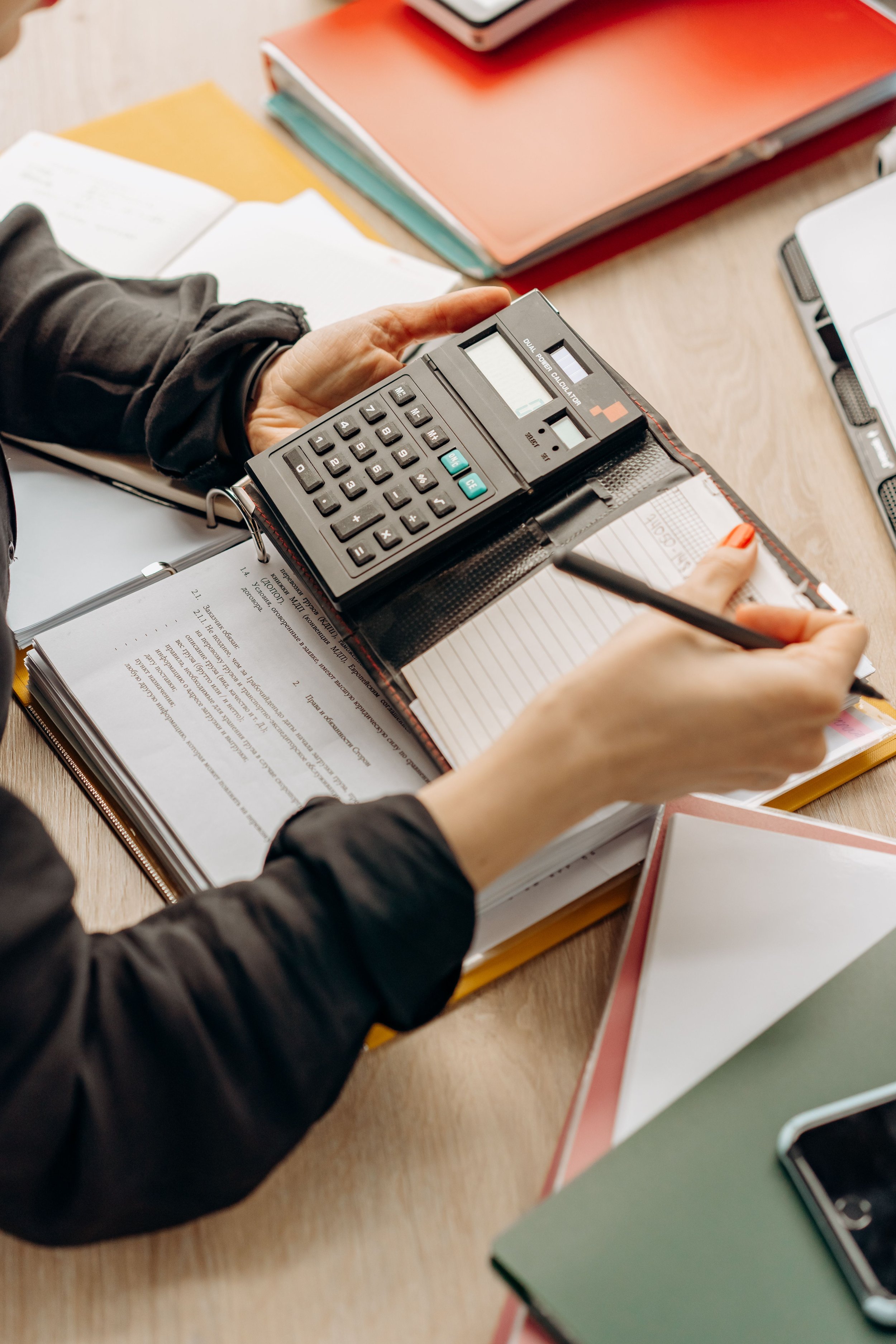Person holding a calculator while taking notes on a notepad, surrounded by notebooks, loose papers, and a mobile phone on a desk.