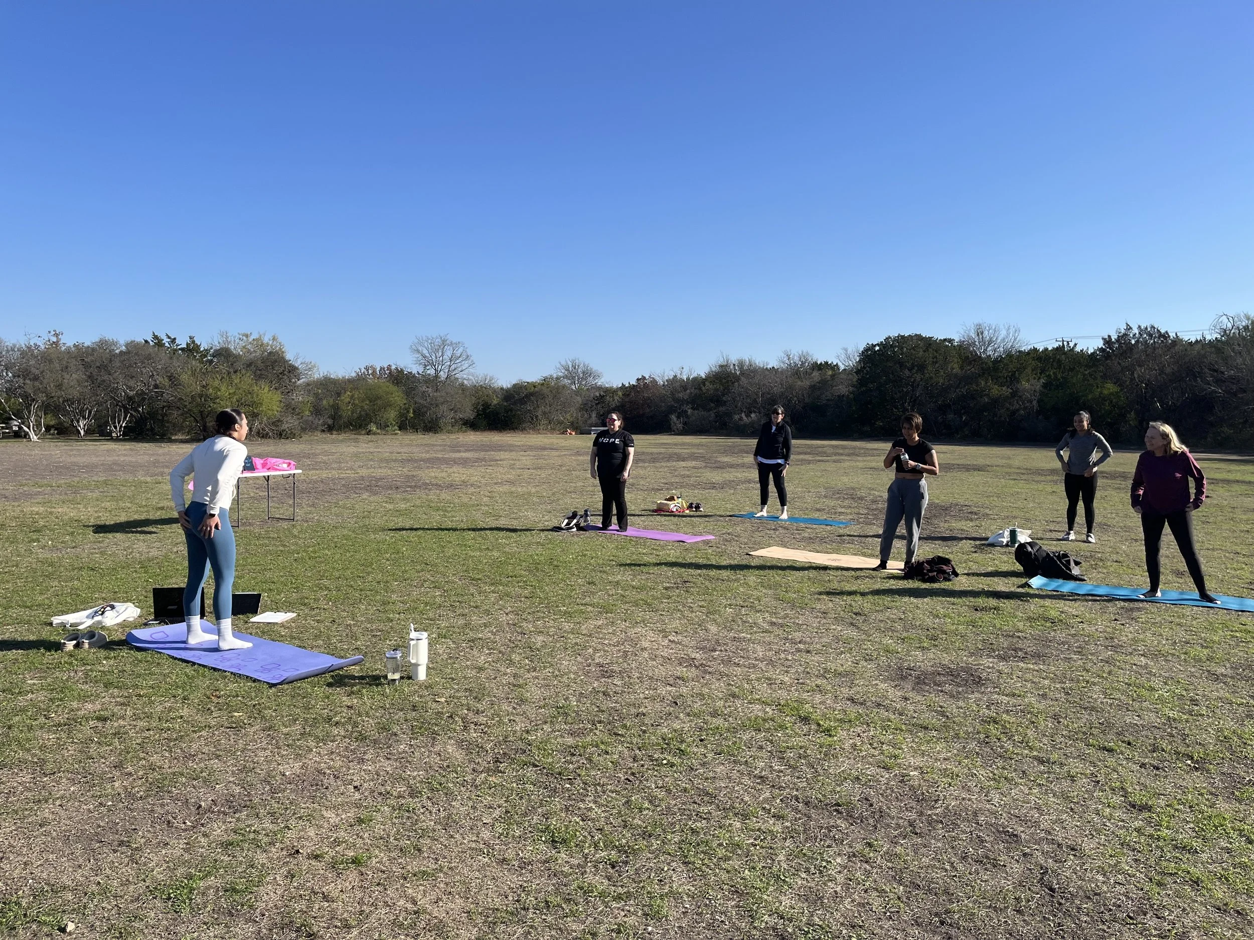 A group of people participating in an outdoor yoga or fitness class on a grassy field on a clear, sunny day. There are mats and water bottles on the ground, and trees in the background.