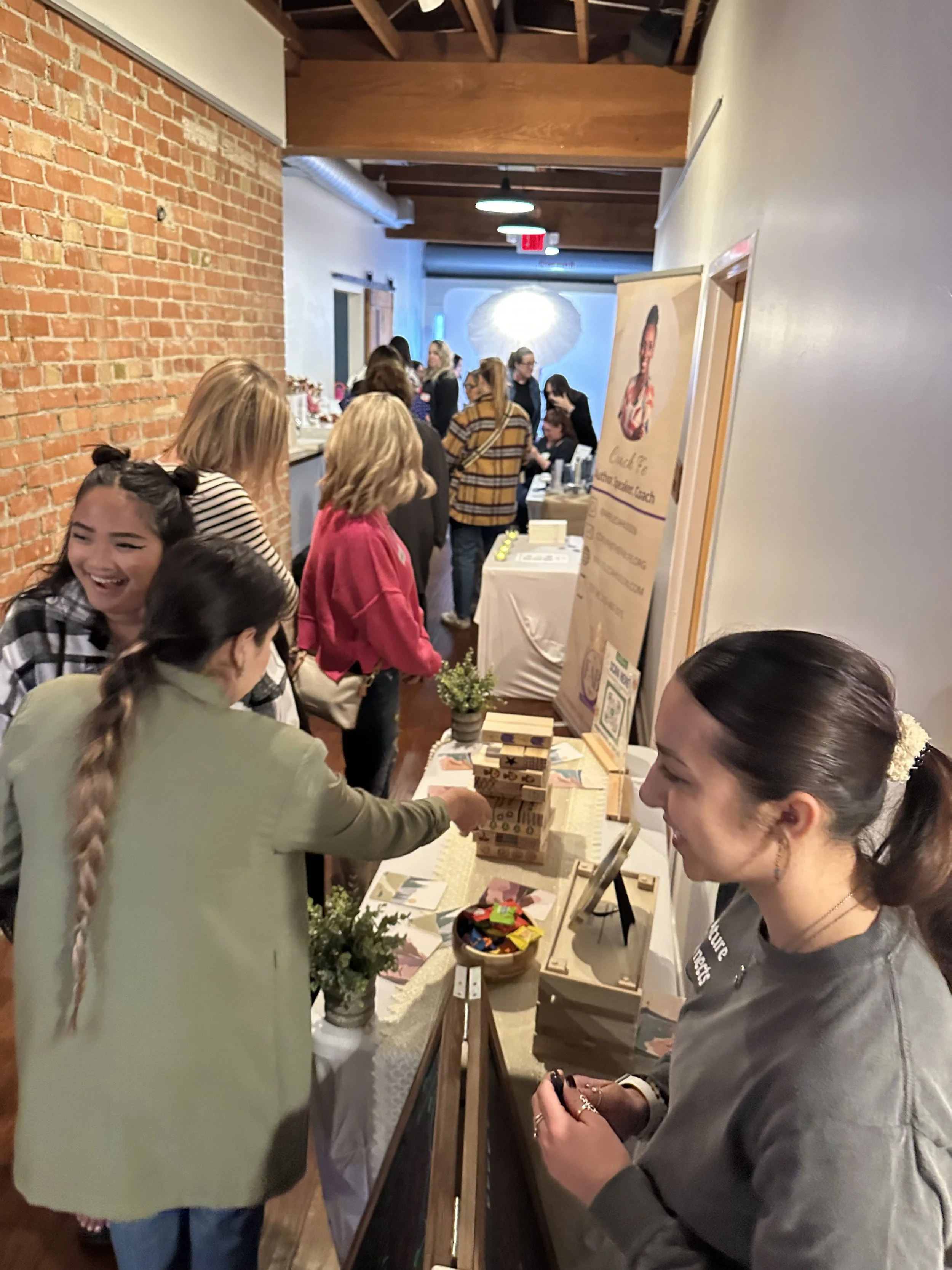 People gathered around tables at an indoor event in a gallery or similar space with brick and white walls, some looking at displays, with lighting fixtures and an exposed ceiling.